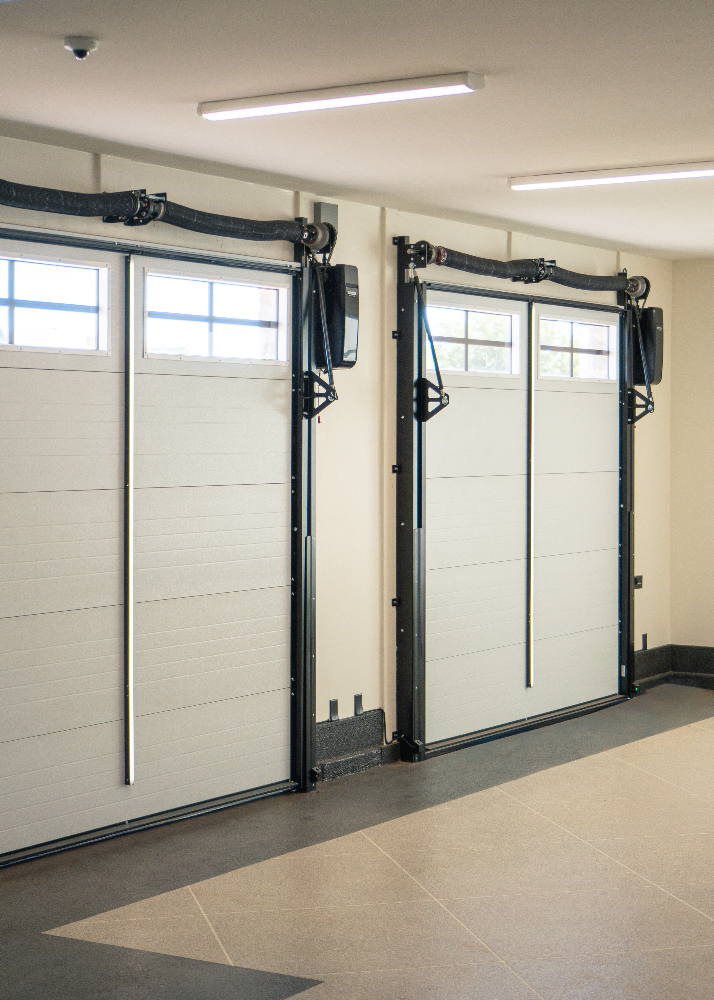 Two white garage doors with small windows at the top, surrounded by black metallic frames, situated inside a well-lit room with gray flooring and ceiling lights.