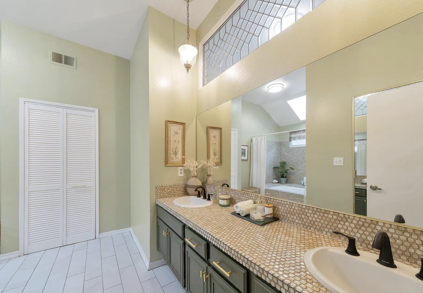 Bathroom with two sinks, large mirror, green cabinets with gold hardware, beige and brown mosaic tile countertop, decorative vases with dried flowers, framed artwork, and a walk-in shower with curtain and a plant.