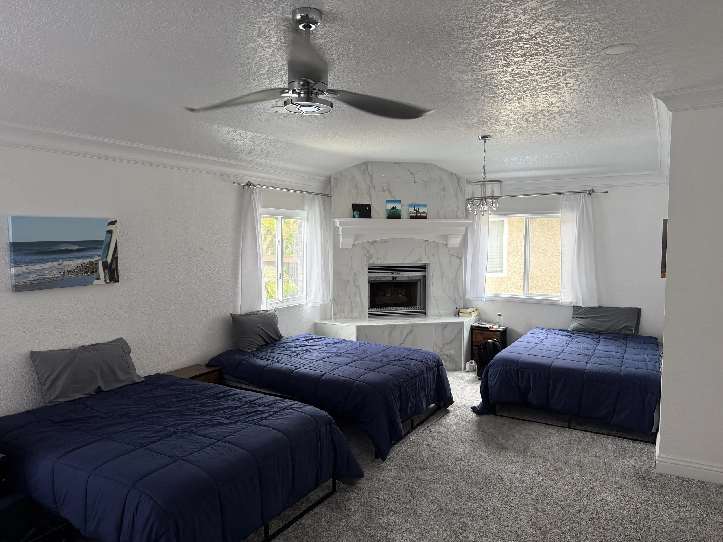 A bedroom with three beds with navy blue bedding, white curtains, a marble fireplace, two windows, and a ceiling fan.