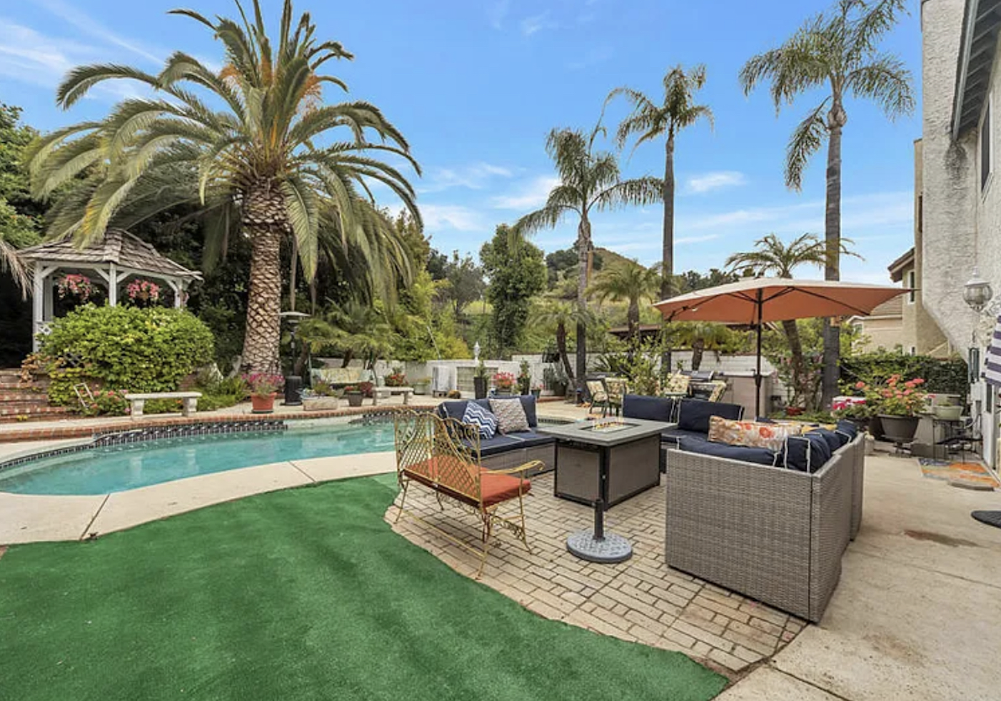 A backyard with a swimming pool, outdoor seating, palm trees, and lush greenery under a blue sky.