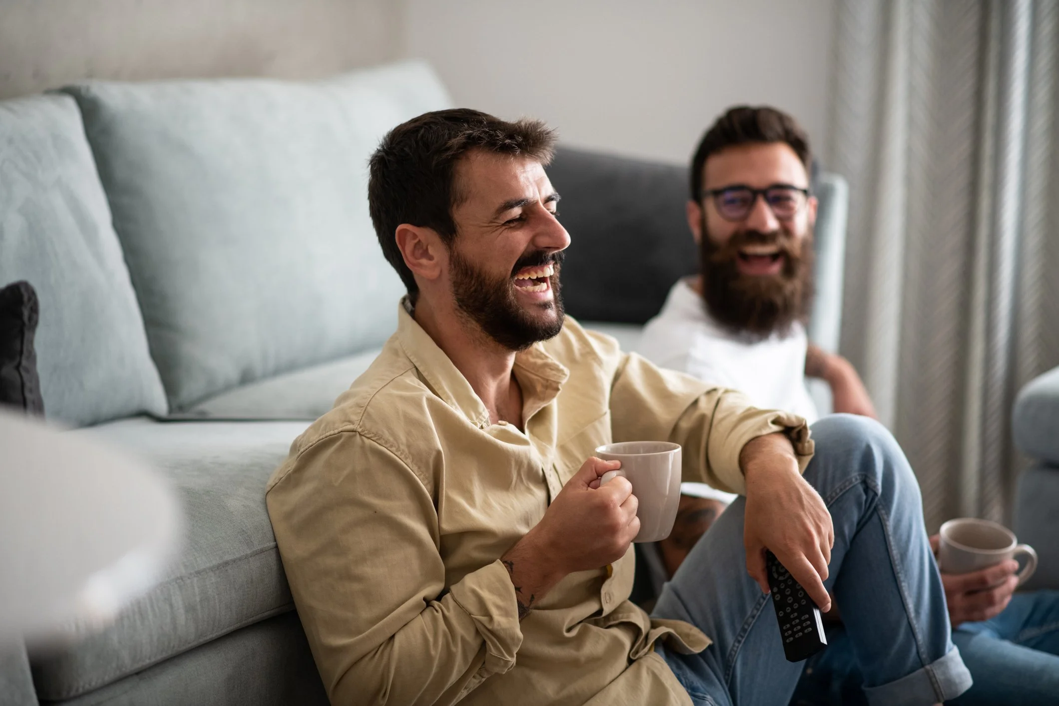 Two men sitting on a couch, smiling and laughing, holding coffee mugs, in a cozy living room.