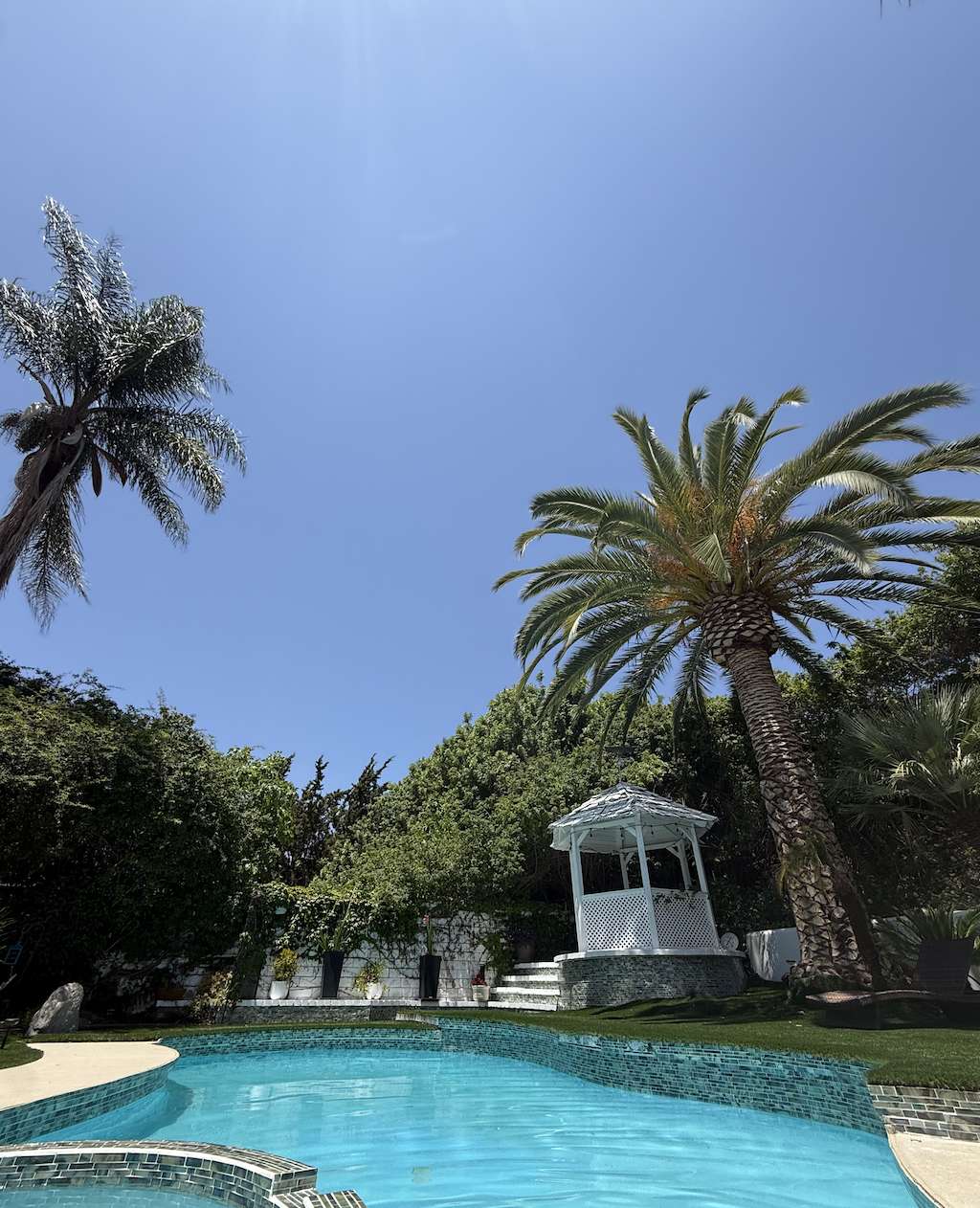 A swimming pool surrounded by tropical palm trees, with a white gazebo on a stone platform and a clear blue sky overhead.