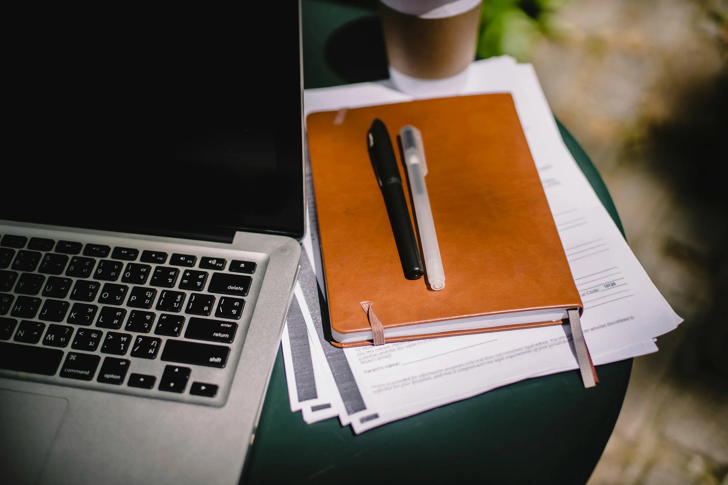 Laptop, orange notebook, black and clear pens, stacks of printed papers, and a brown paper cup on a dark green table outdoors.