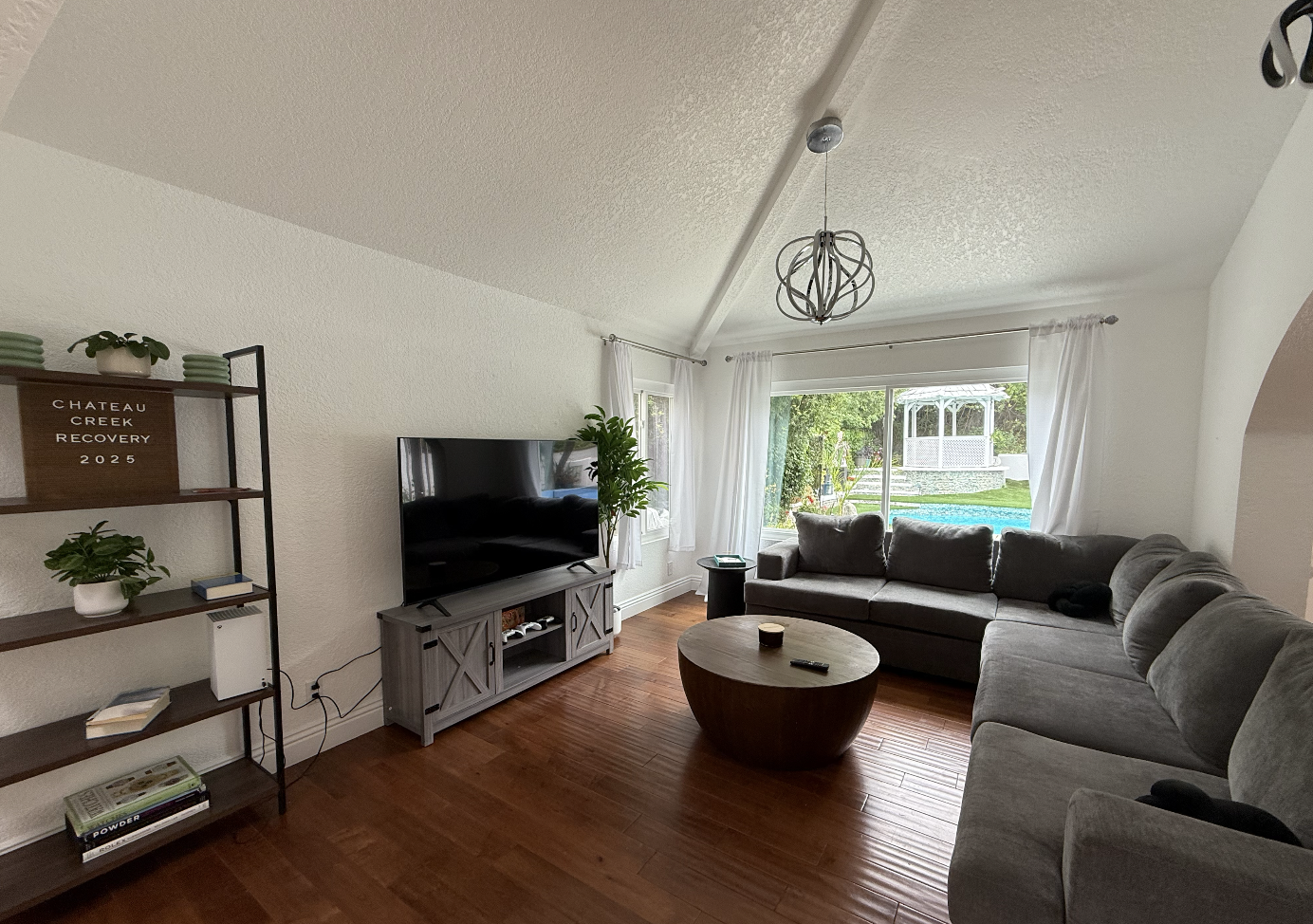 Living room with gray sectional sofa, round wooden coffee table, TV on a gray stand, large window with white curtains, and backyard with swimming pool and gazebo