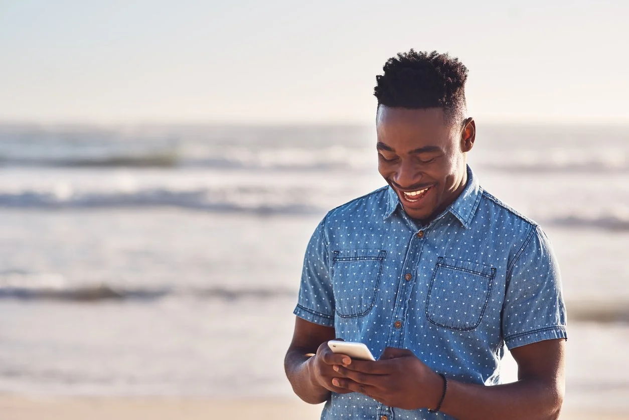 A young man smiling and looking at his phone on the beach with waves in the background.