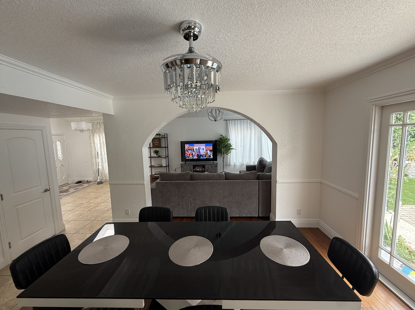 Dining room with a black table, six black chairs, a chandelier, and a view into the living room with a TV, sofa, and plant.
