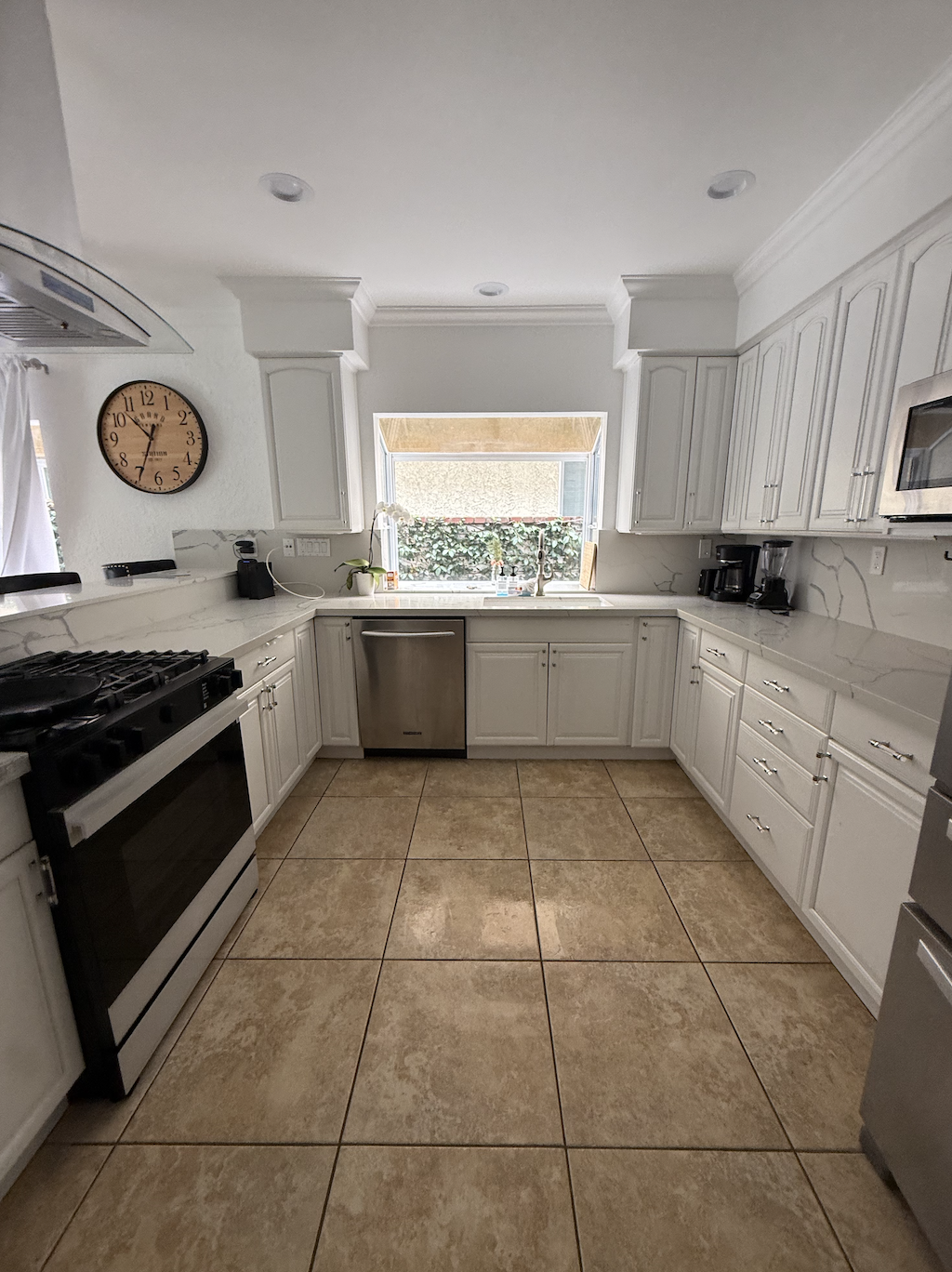 White kitchen with tiled floor, clock on wall, window over sink, stainless steel appliances, and black coffee maker on countertop.