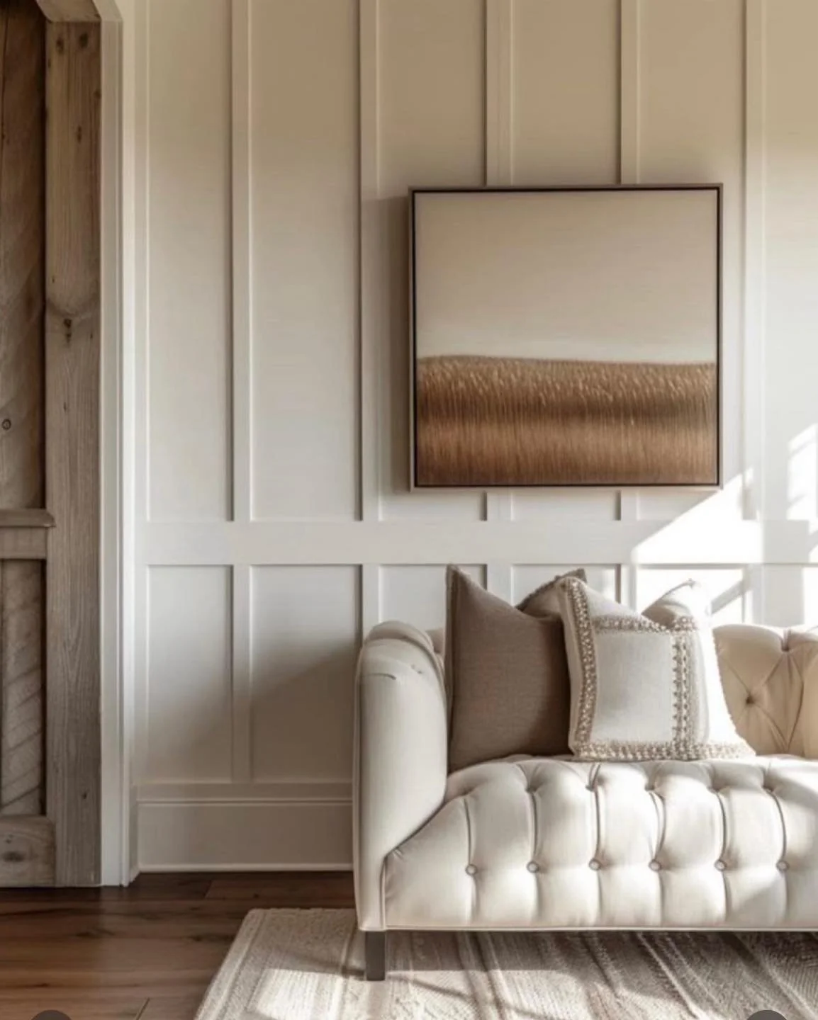 Living room with white paneled wall, framed abstract art, beige tufted sofa with decorative pillows, wooden floor, beige rug, and sunlight streaming in.