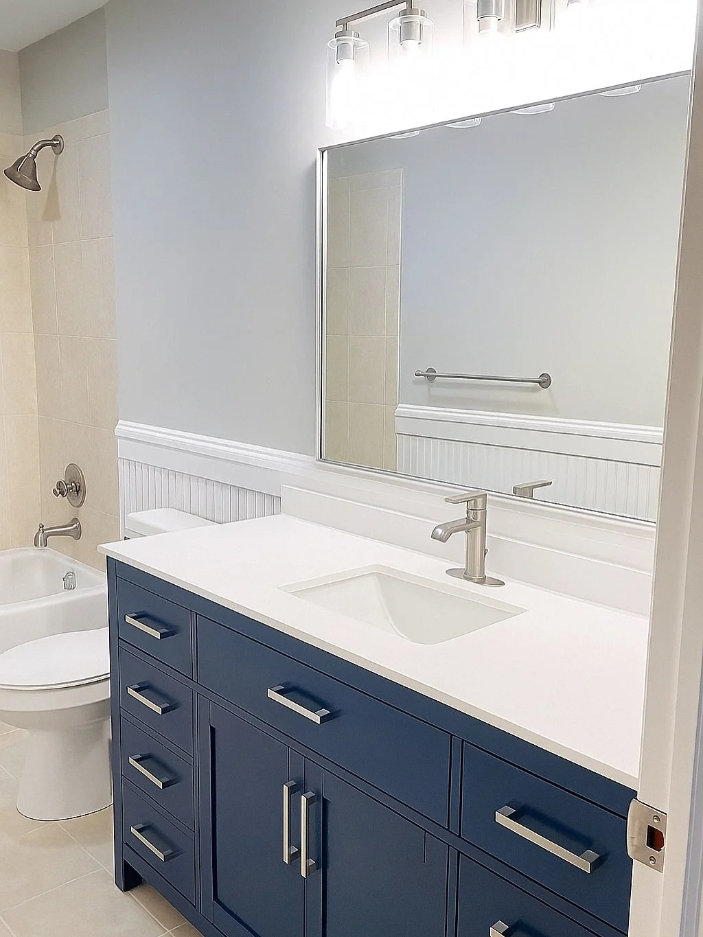 Bathroom with a blue vanity, white countertop, and a large mirror. There is a shower with a chrome showerhead and a bathtub with a chrome faucet. Beige tile on the shower wall, and a towel bar on the wall.