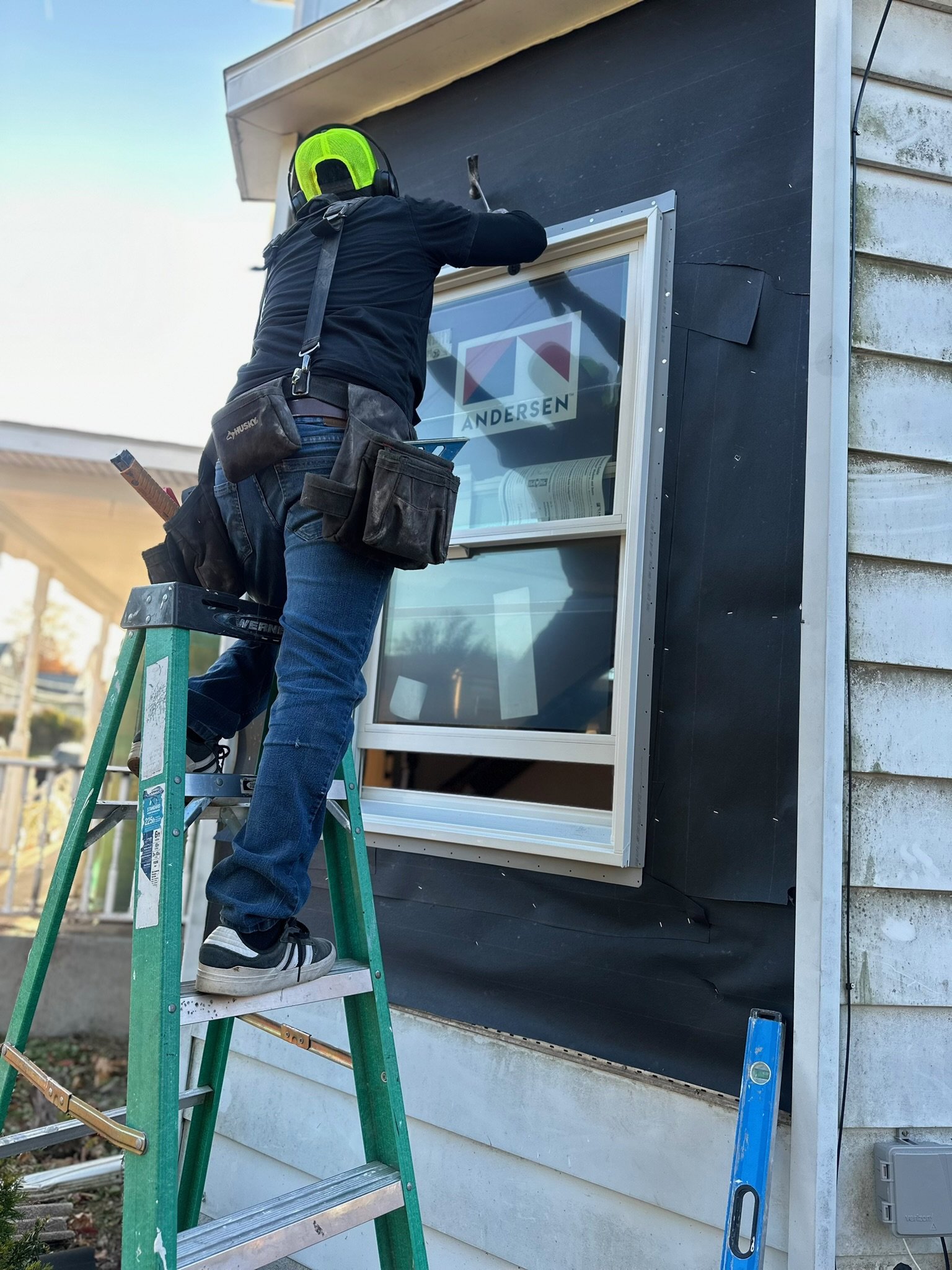 A person standing on a green ladder installing or repairing a window exterior on a house, with tools and a tool pouch around their waist, wearing a black shirt, jeans, and a neon yellow safety helmet.