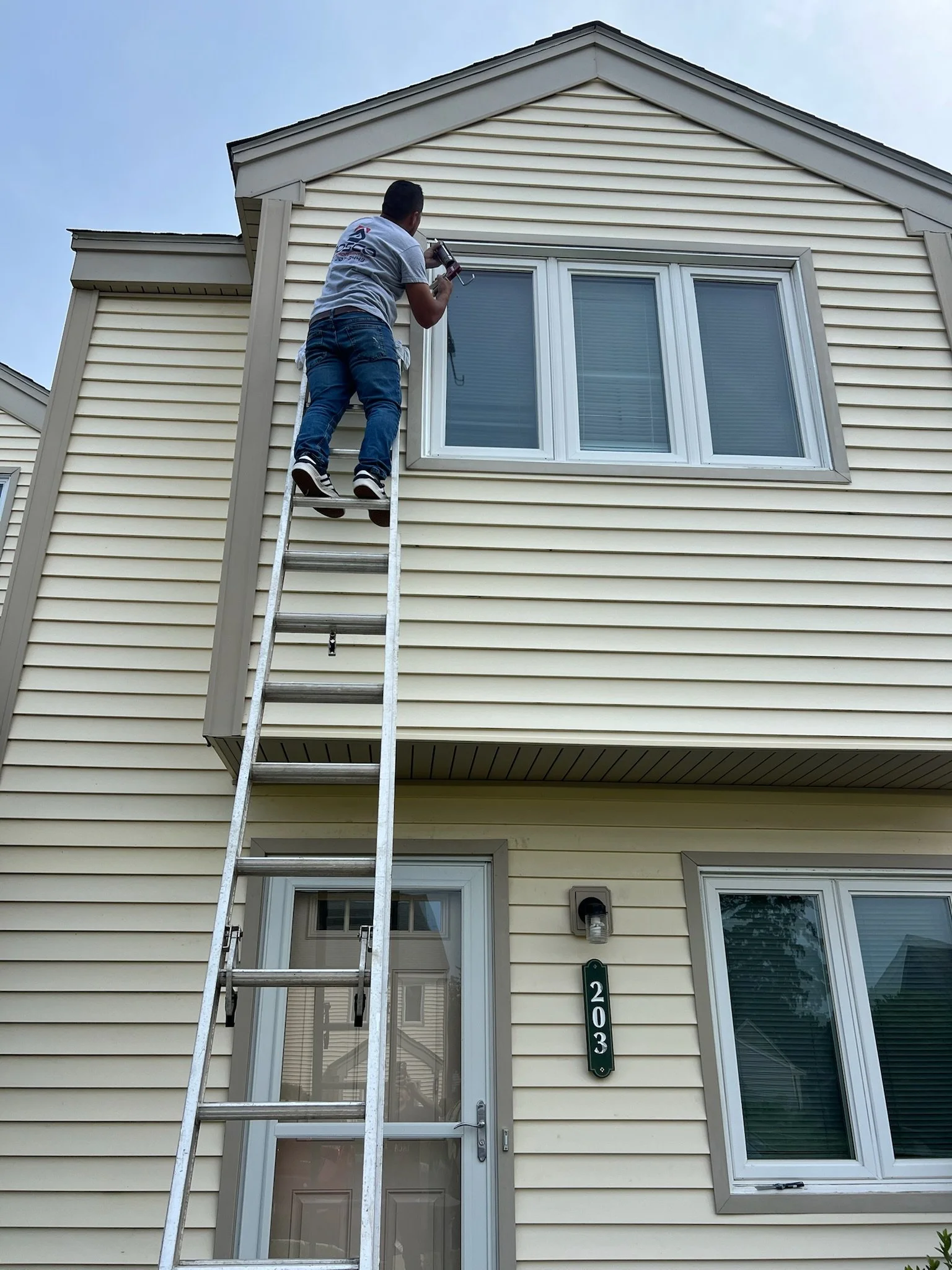 A man on a ladder cleaning or repairing a window on the second story of a yellow house with beige trim.