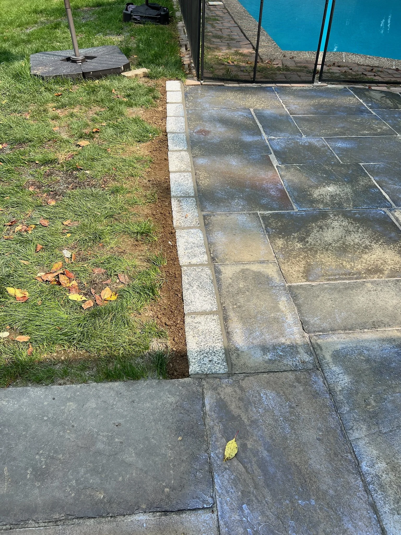 A pool area with a stone patio, a glass safety fence, and a patch of grass with fallen leaves. A large umbrella base is visible on the grass.