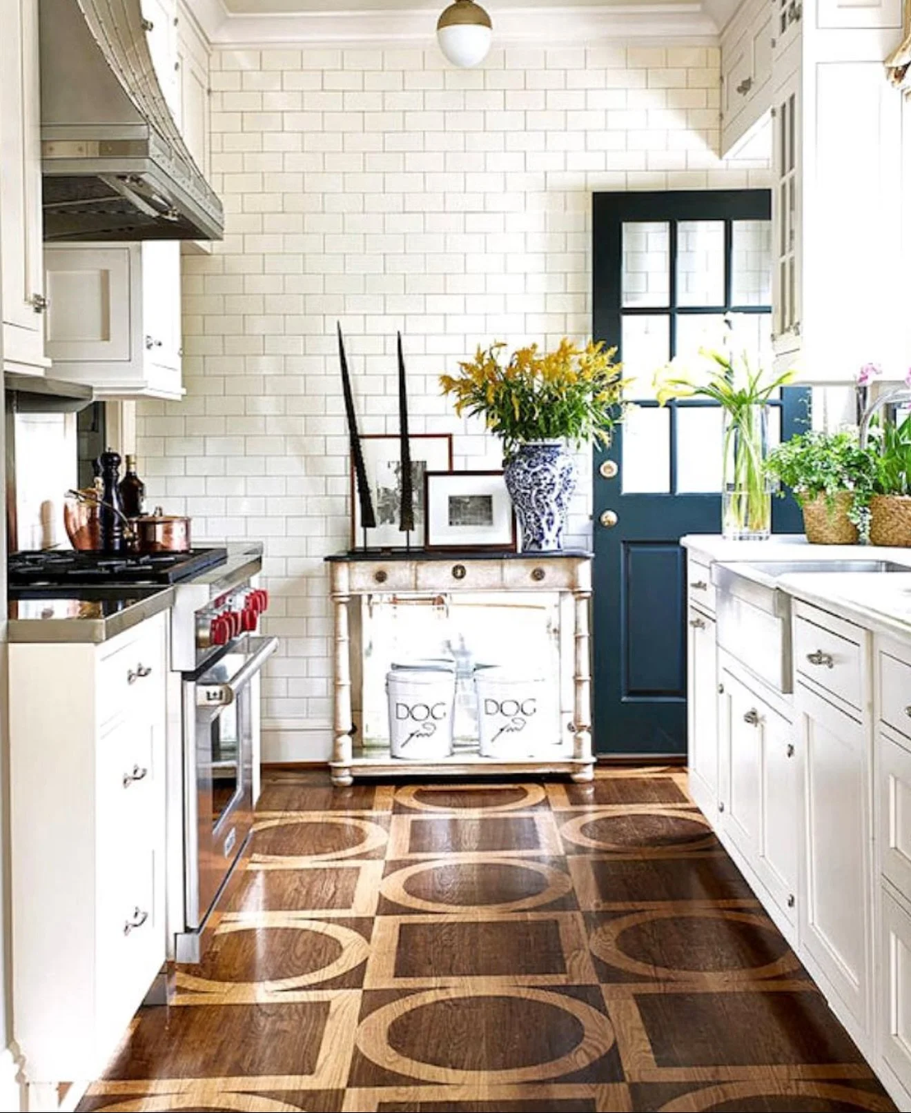 A cozy kitchen with white cabinets, a white brick wall, and hardwood flooring featuring a geometric pattern. The kitchen has potted plants near a window and a dark blue door. There is a small white distressed table with framed pictures and flowers on