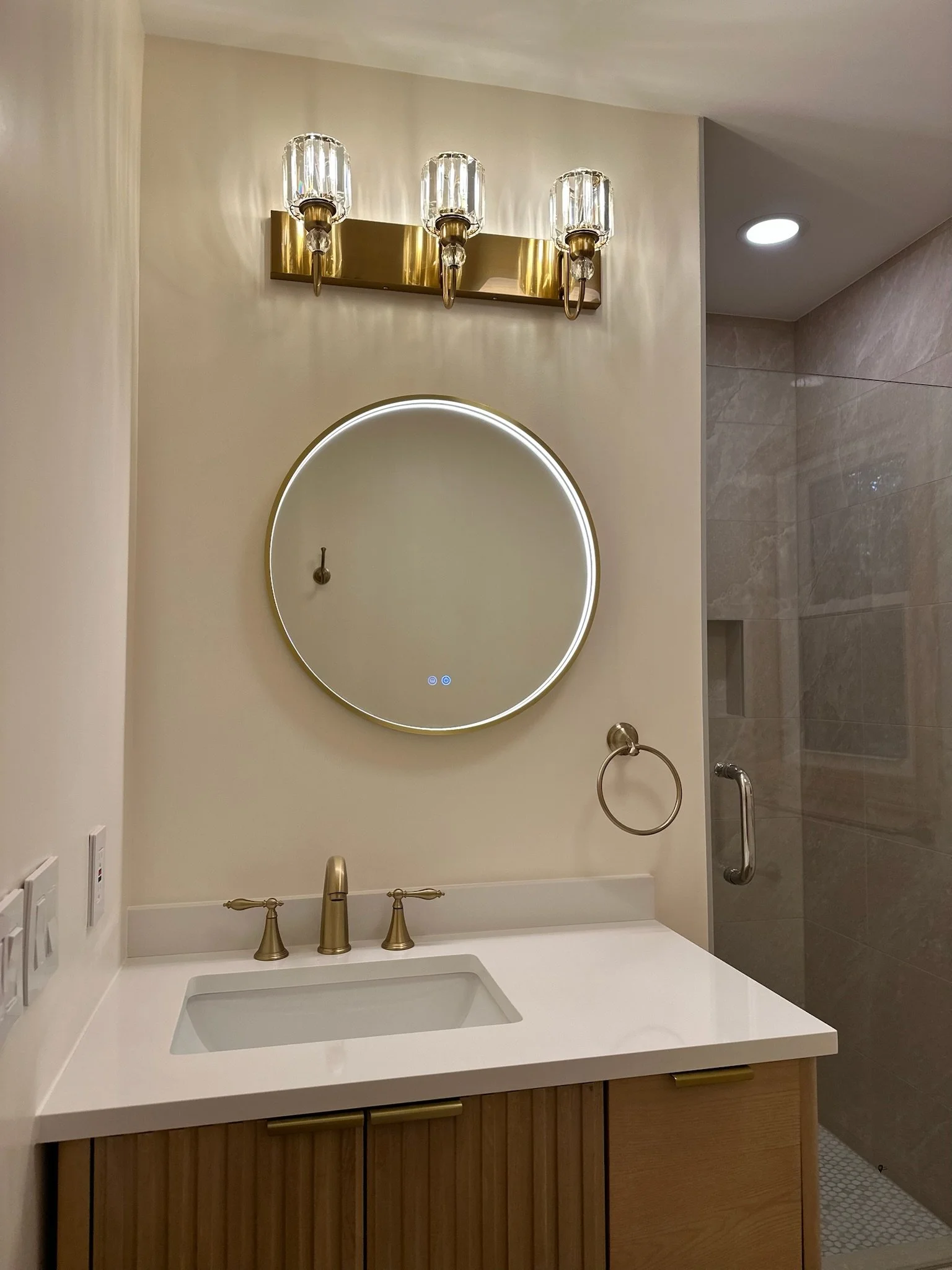 Bathroom vanity with a round illuminated mirror, gold fixtures, a wooden cabinet, a gold towel ring, and a shower area with a glass door.