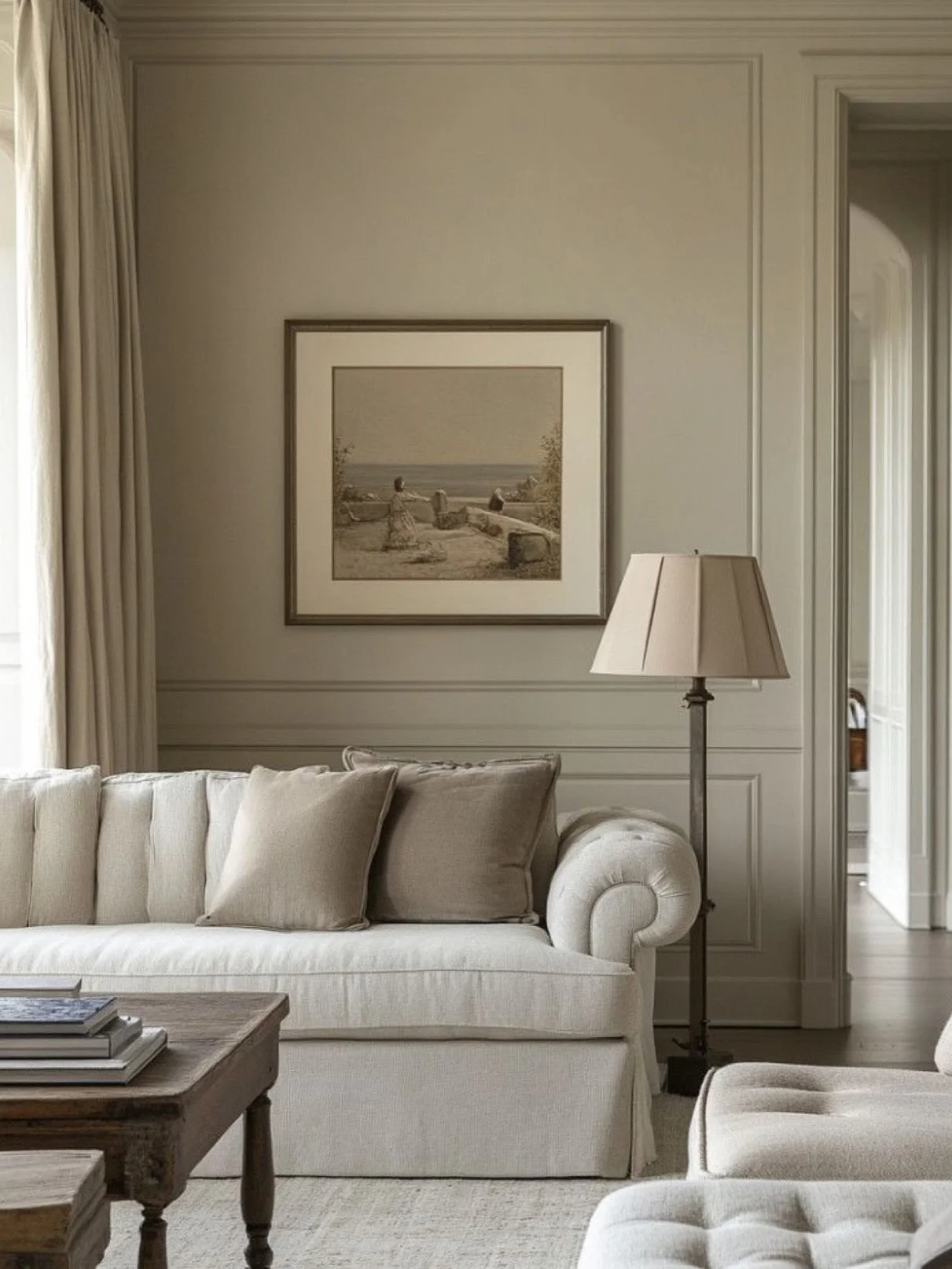 A cozy living room corner with a white sofa and beige pillows, a wooden coffee table with books, a floor lamp, a framed picture of a beach scene on the wall, and a window with cream curtains.