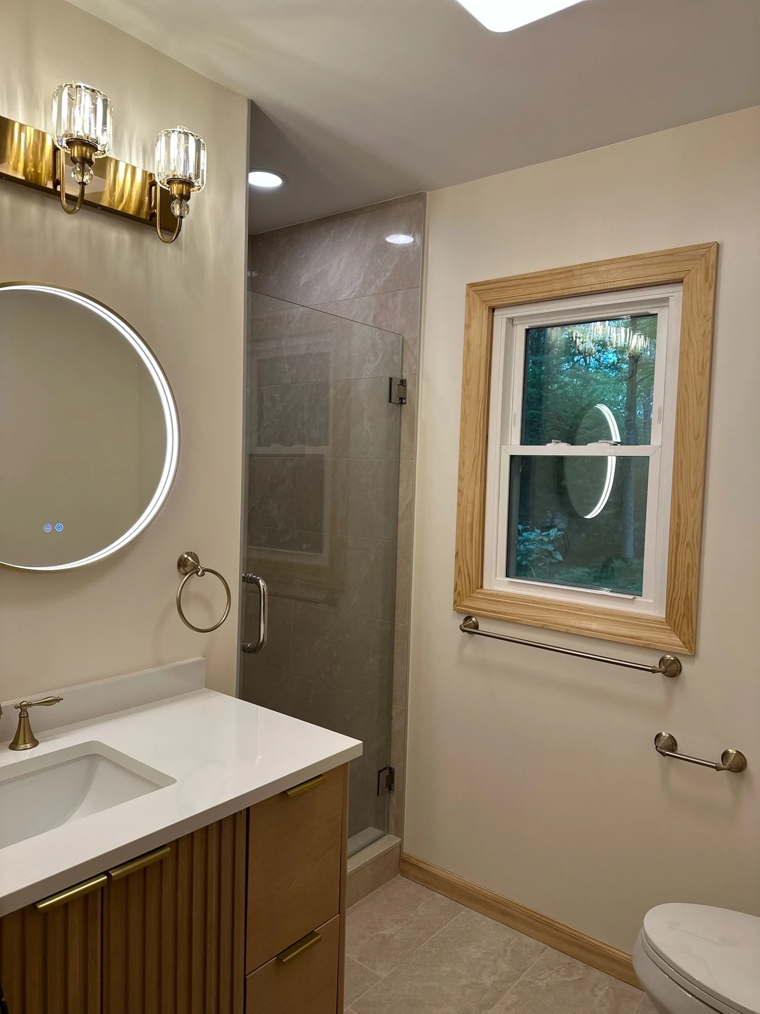 Bathroom with vanity, round mirror, wall light fixture, window with wooden frame, shower with glass door, and toilet.