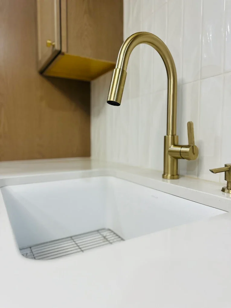 Close-up of a white kitchen sink with a metallic gold faucet and handles, mounted on a white marble or quartz countertop, with a wooden cabinet and beige wall in the background.