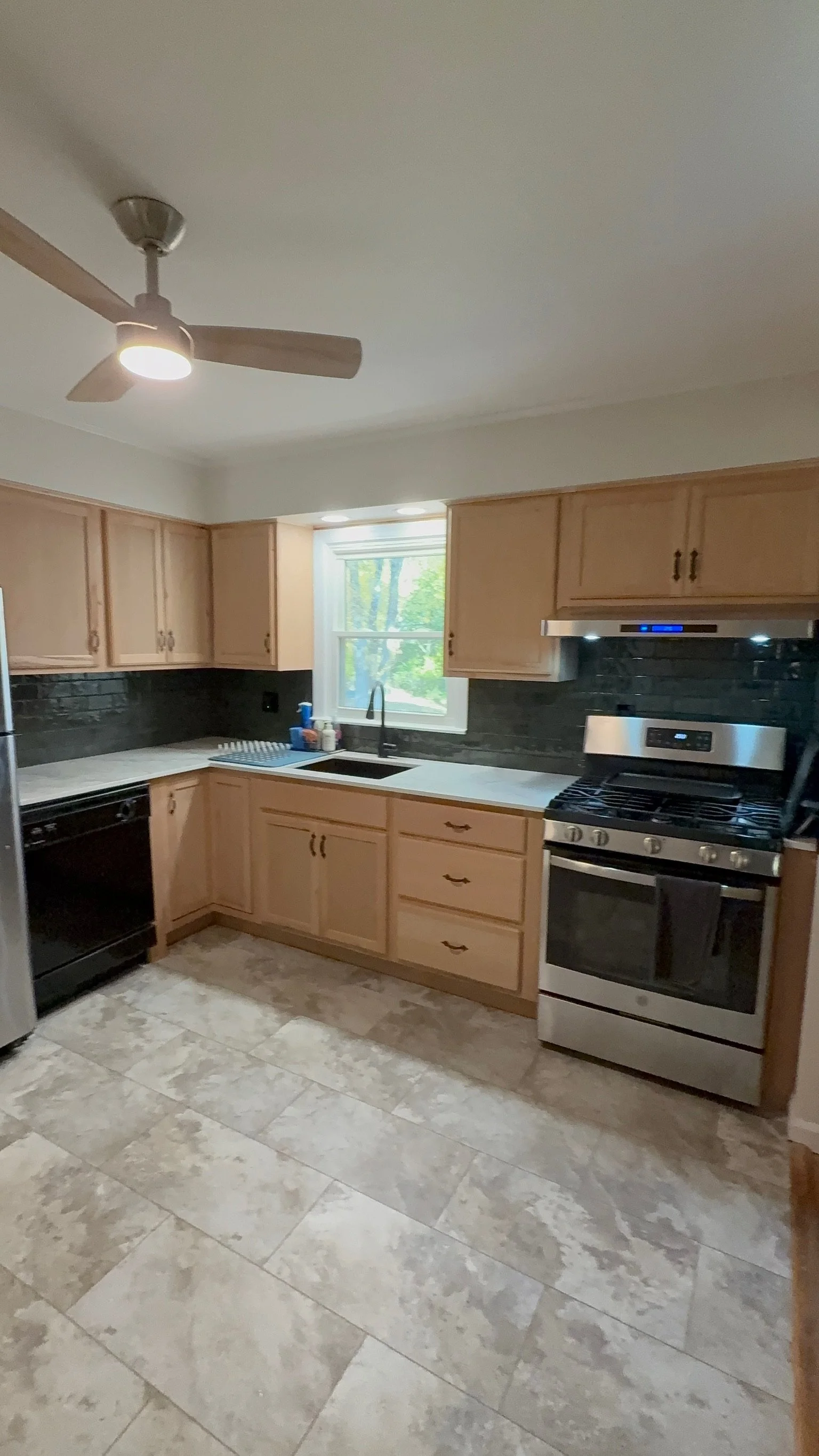 Kitchen with wood cabinets, black backsplash, stainless steel stove, black dishwasher, window above the sink, ceiling fan with light, tiled floor.