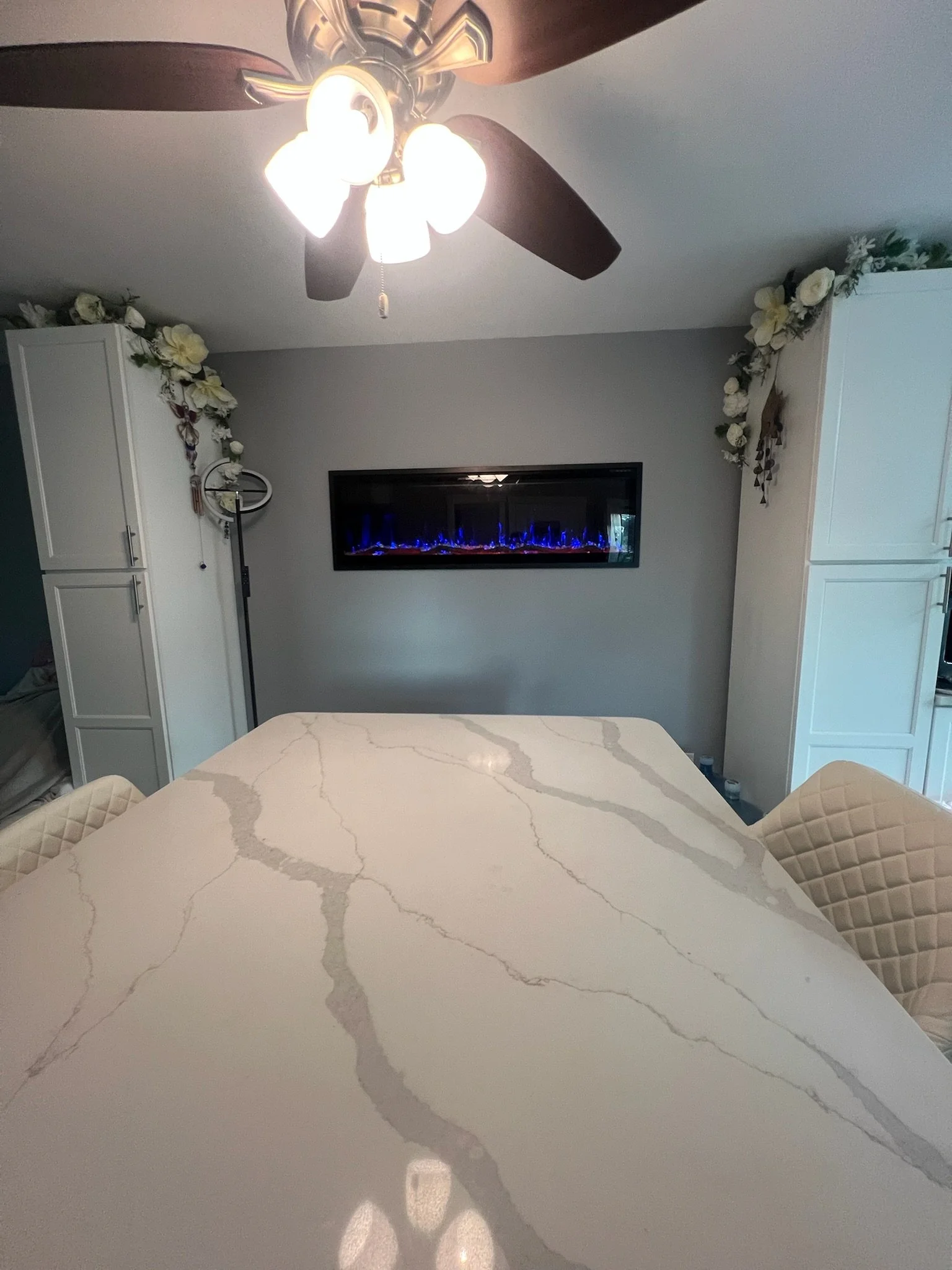 A dining table with a white marble top featuring gray veining, surrounded by quilted cream chairs. The background includes a gray wall with a modern electric fireplace displaying blue flames, white cabinets with floral decorations, and a ceiling fan 