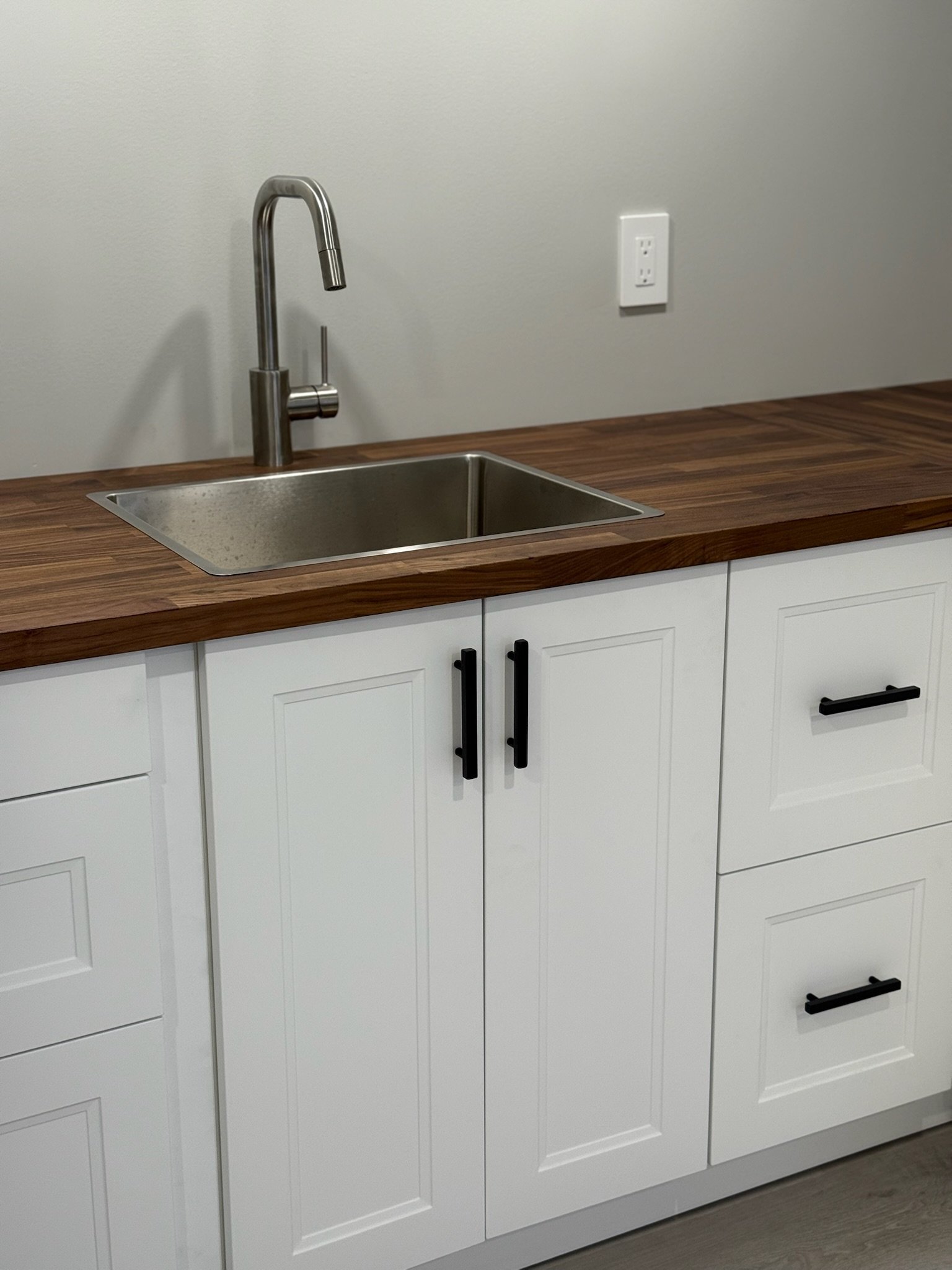 A modern kitchen sink with a stainless steel basin, a tall curved faucet, white cabinetry with black handles, and a wooden countertop. A white electrical outlet is on the gray wall behind the sink.