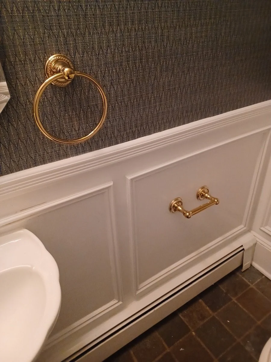 Close-up of a bathroom wall with a gold towel ring and a gold toilet paper holder, part of a toilet visible at the bottom left, dark tiled floor.