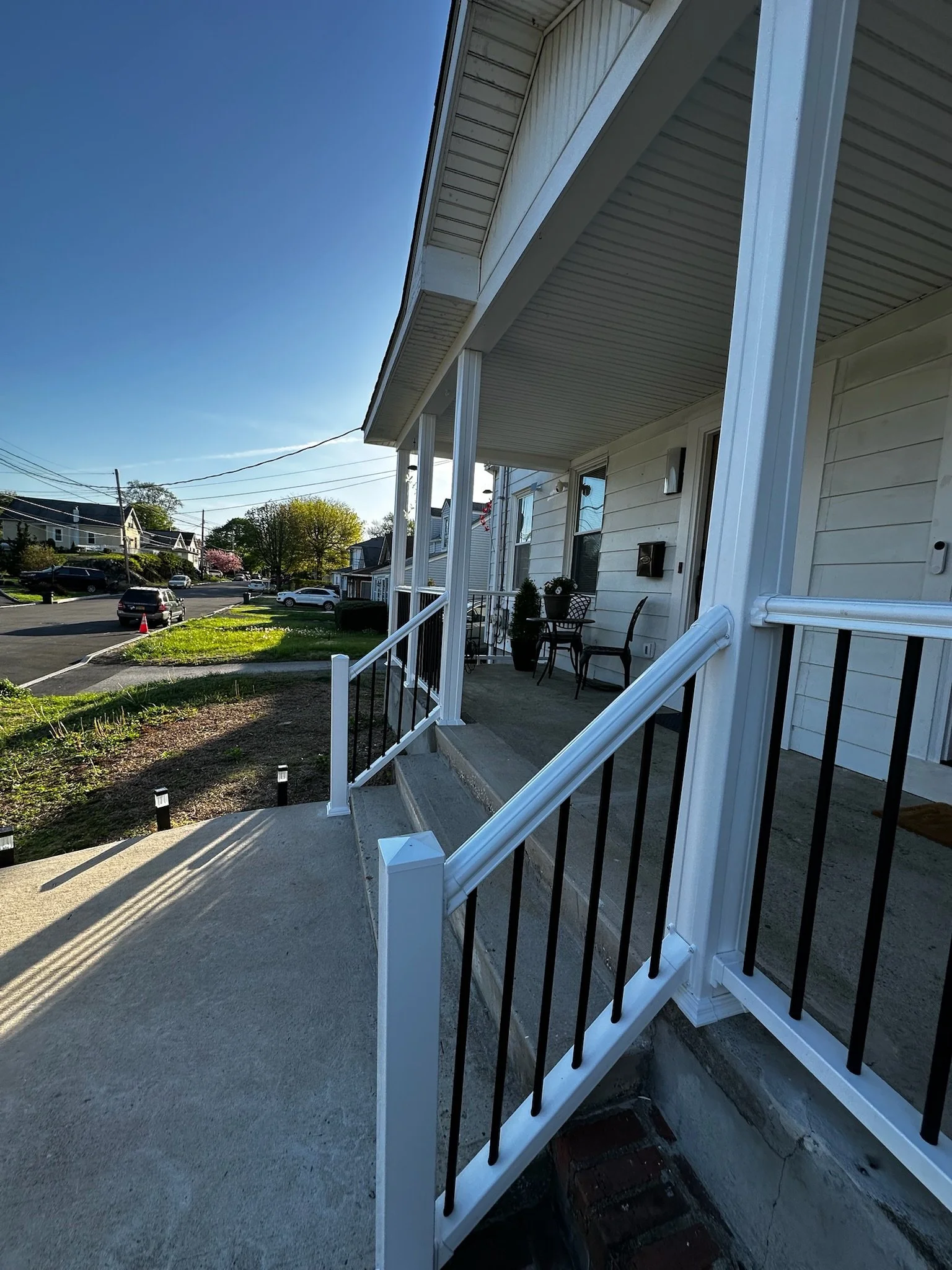 Front porch of a white house with a small seating area, black railing, and steps leading down to the sidewalk, with a residential neighborhood and parked cars in the background under a clear blue sky.