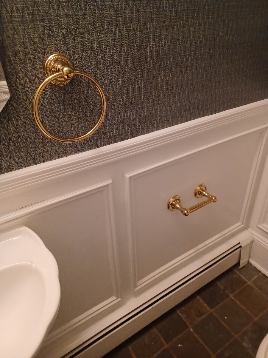 Close-up of a bathroom wall with a gold towel ring and a gold toilet paper holder, white wainscoting, dark patterned wallpaper, and tile flooring.
