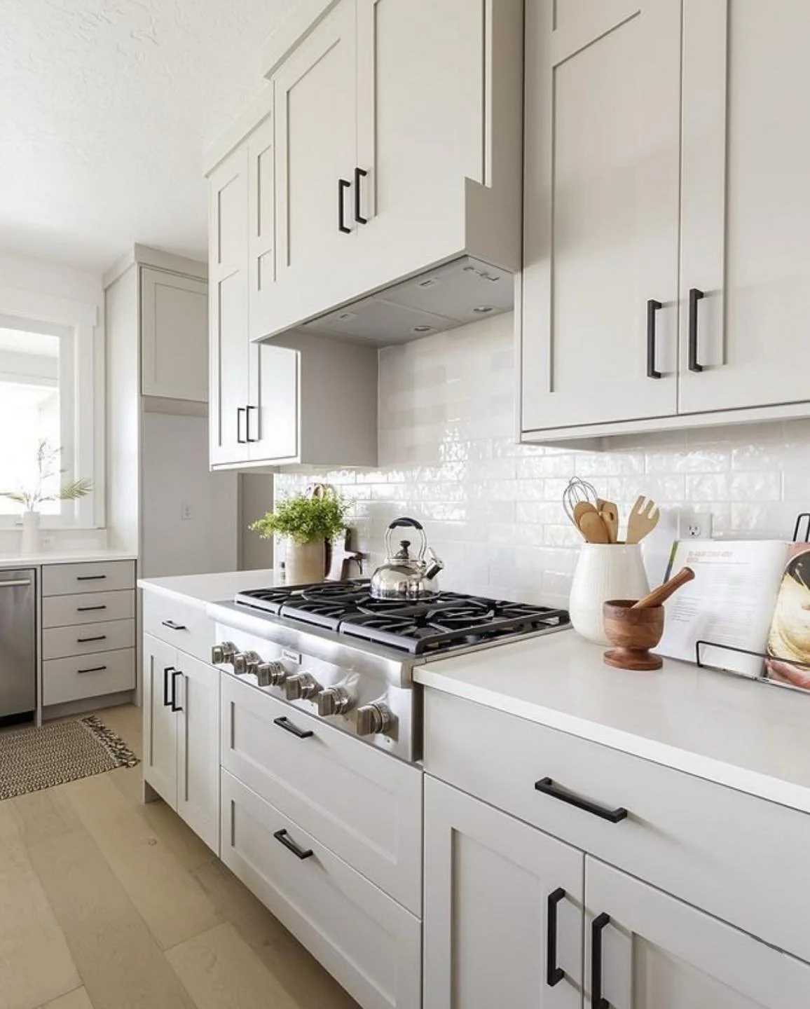 Modern kitchen with white cabinetry, black handles, and a stainless steel stove with a tea kettle. Countertop has a plant, utensils in a container, a wooden mortar and pestle, and a cookbook.