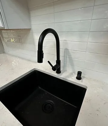 Black kitchen sink with a tall curved black faucet and separate black control knob, set in a white marble countertop against a white tiled wall.
