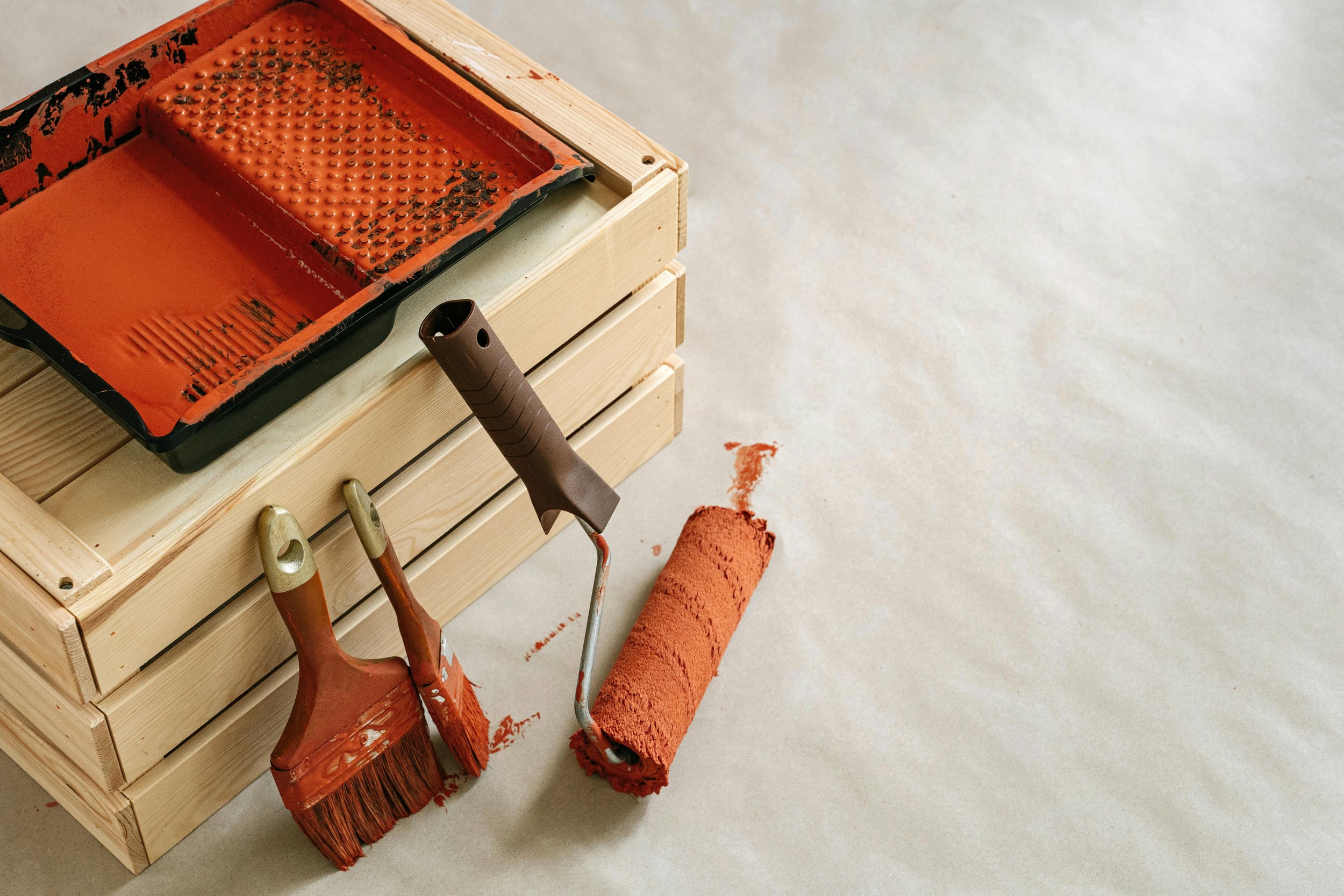 Painting supplies including a paint roller, paintbrush, and paint tray with orange paint, set on a light-colored floor next to a wooden box.