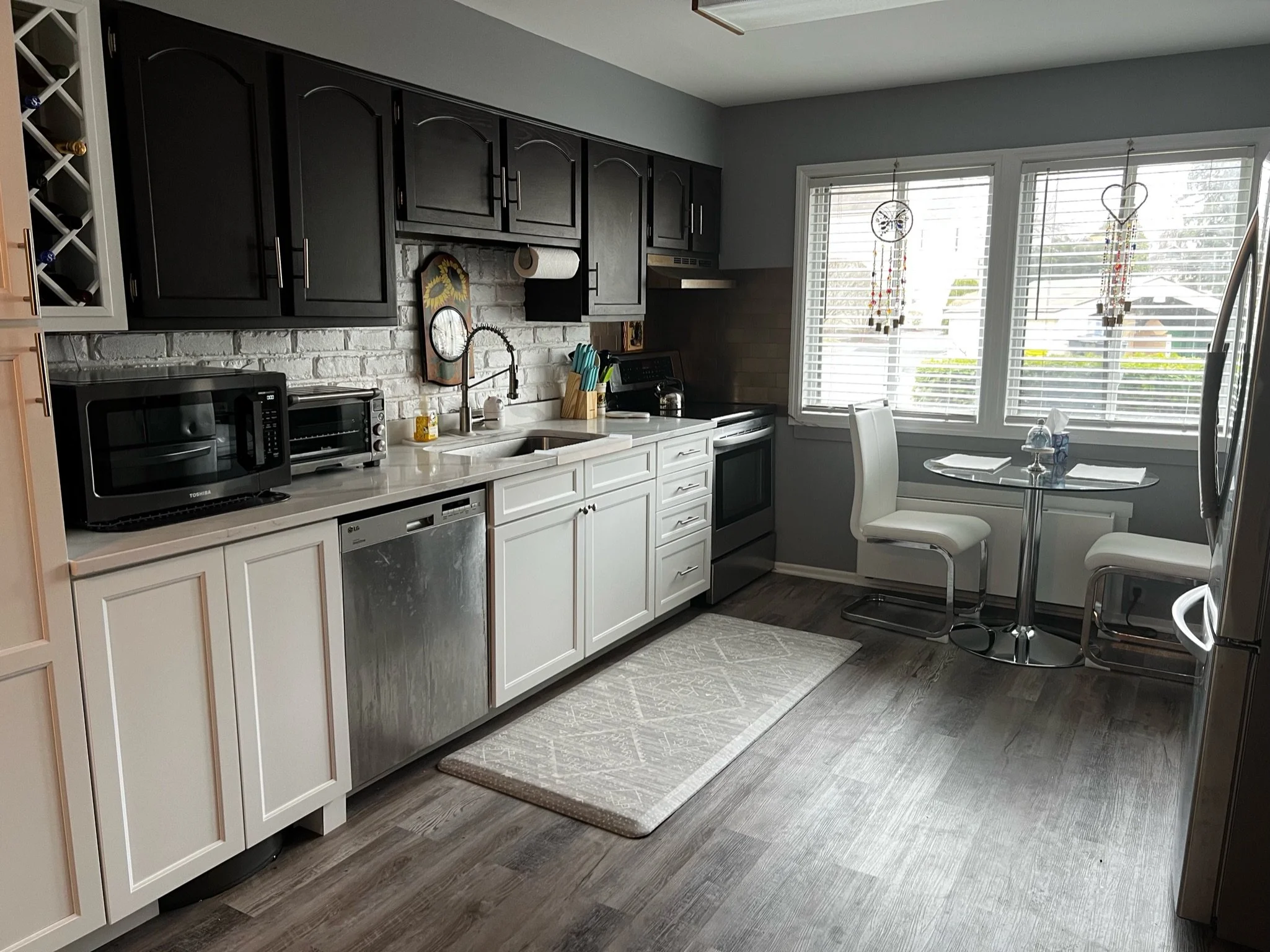 Kitchen with black upper cabinets, white lower cabinets, and a white countertop, featuring small appliances like a microwave, toaster oven, and a dishwasher, with a small dining area near windows with blinds and hanging dreamcatchers.