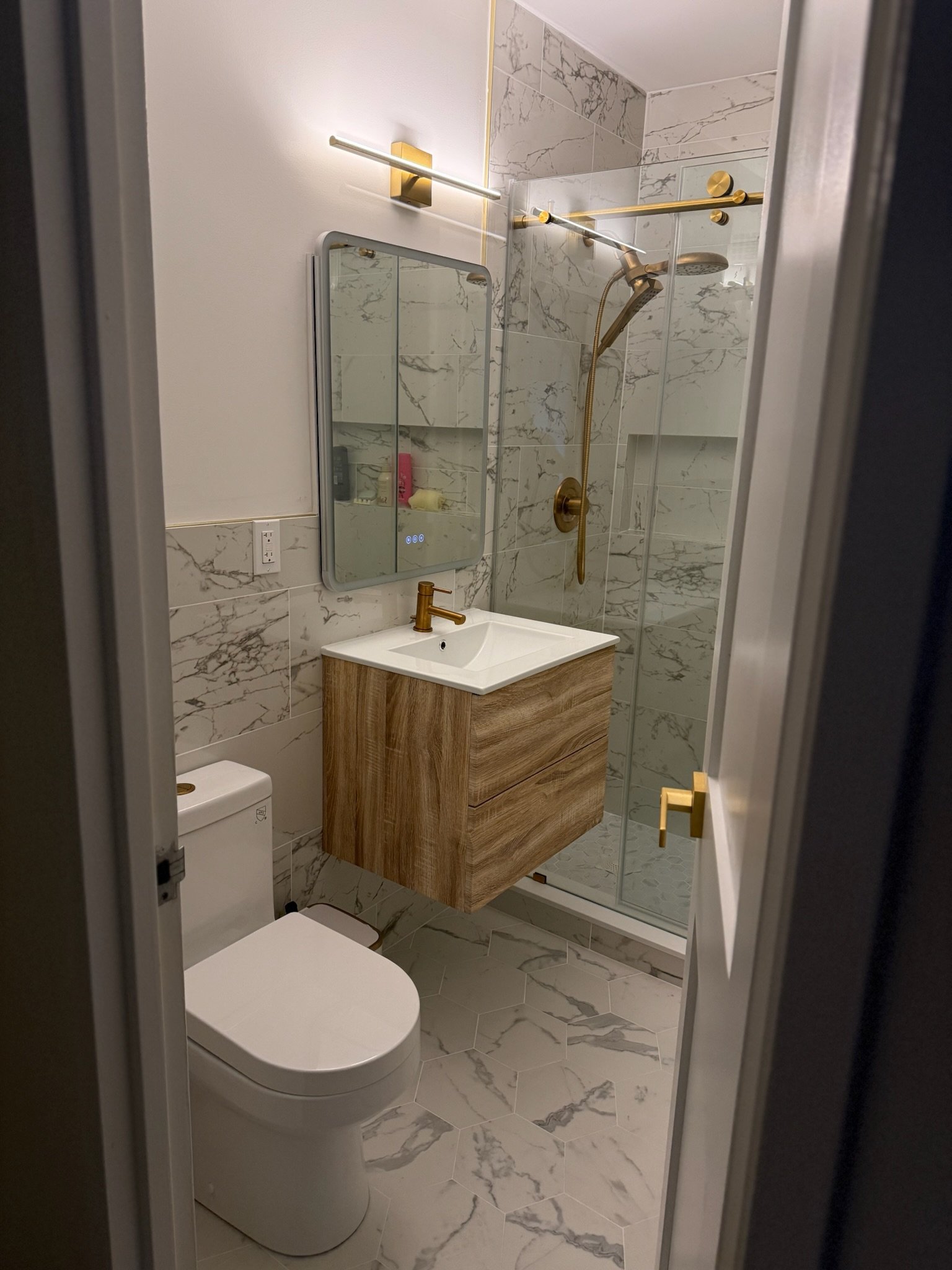 Small modern bathroom with marble tile walls and floor, a wall-mounted wooden vanity with white sink and gold faucet, mirror, toilet, and glass shower with gold fixtures.