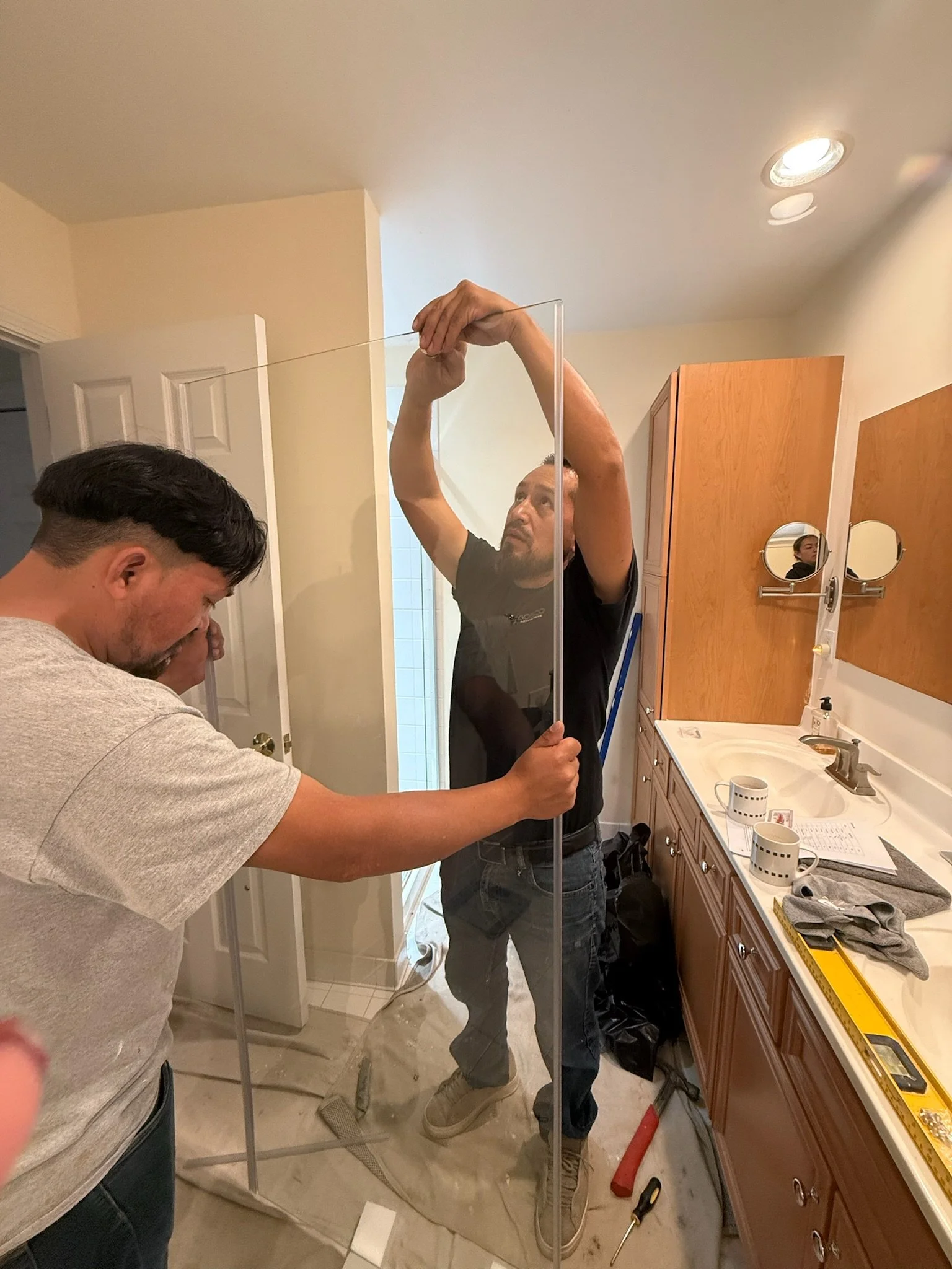 Two men installing a clear glass shower door in a bathroom. One man holds the glass panel in place while the other adjusts the top.