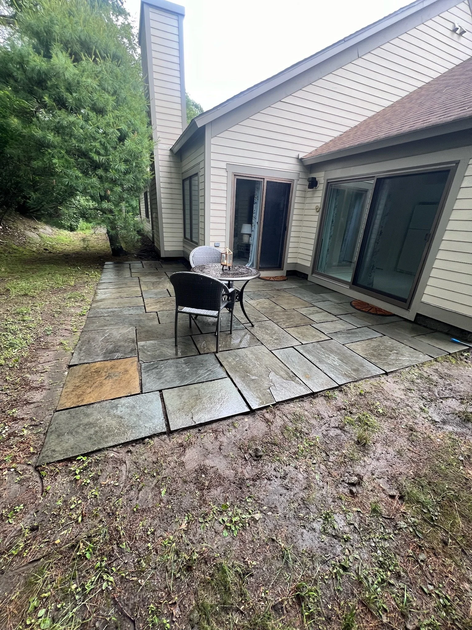 Backyard patio with stone pavers, outdoor table and chairs, sliding glass doors, and surrounded by mud and greenery.