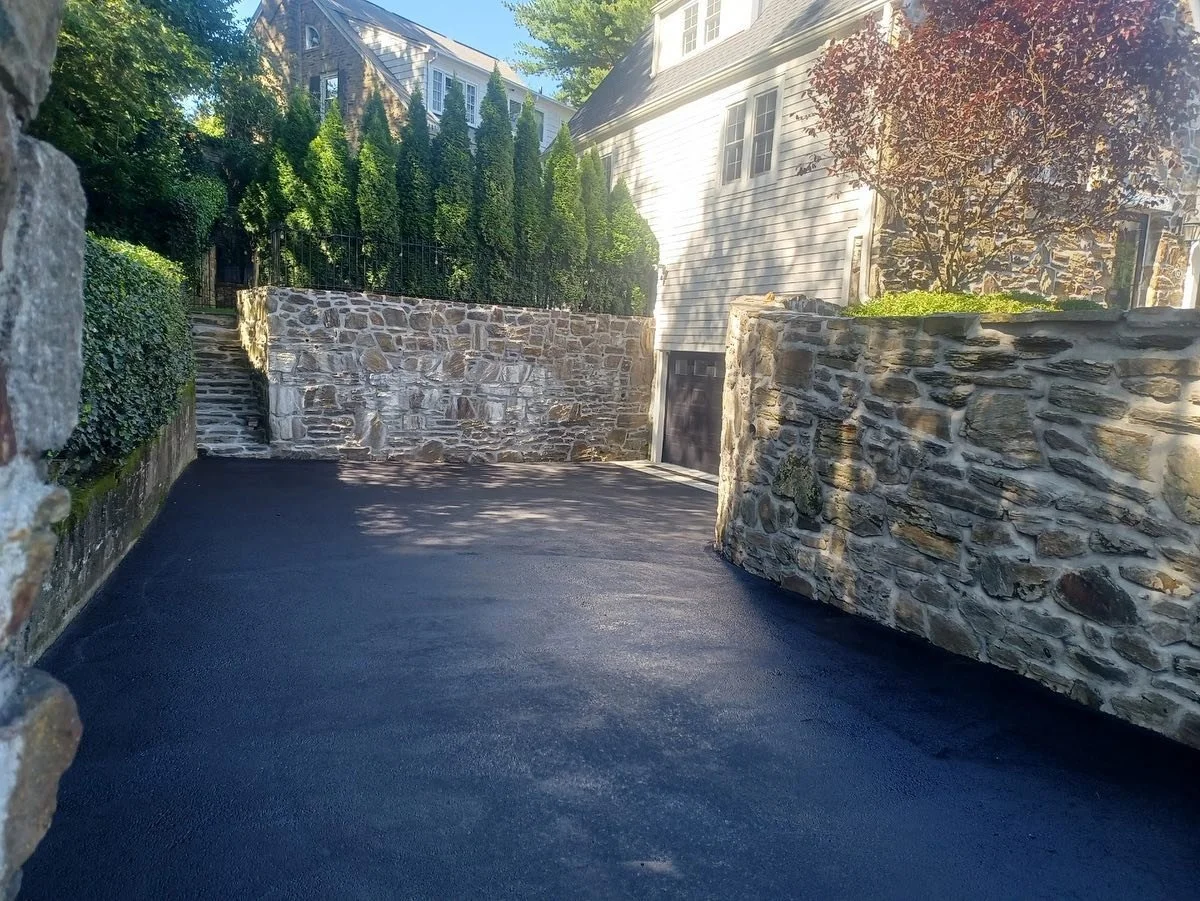 A paved driveway with new black asphalt, surrounded by stone walls and green shrubbery in a residential area.