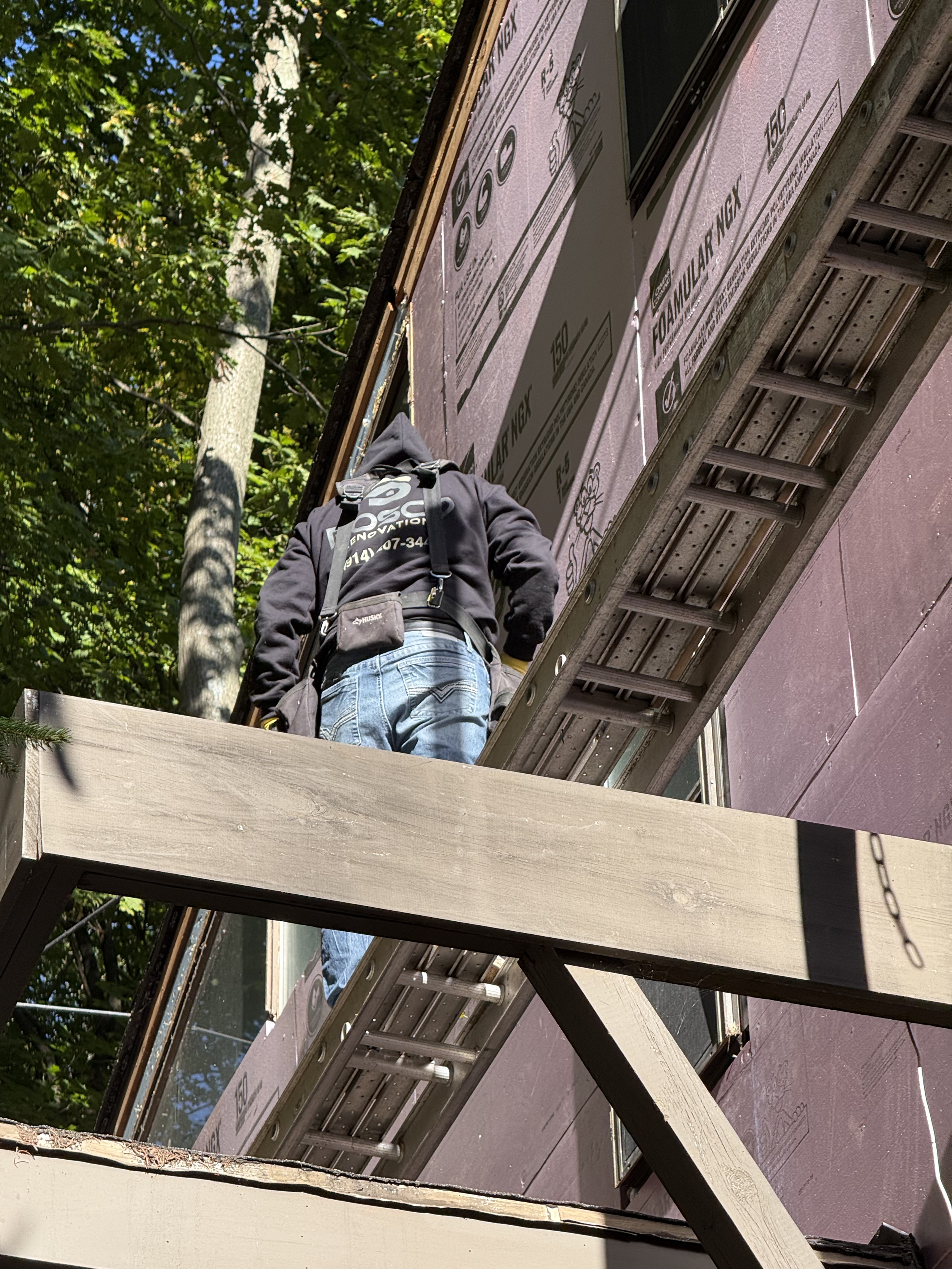 A construction worker on a ladder working on the exterior of a house during daytime. The house is partially covered with pink insulation board, and greenery is visible in the background.