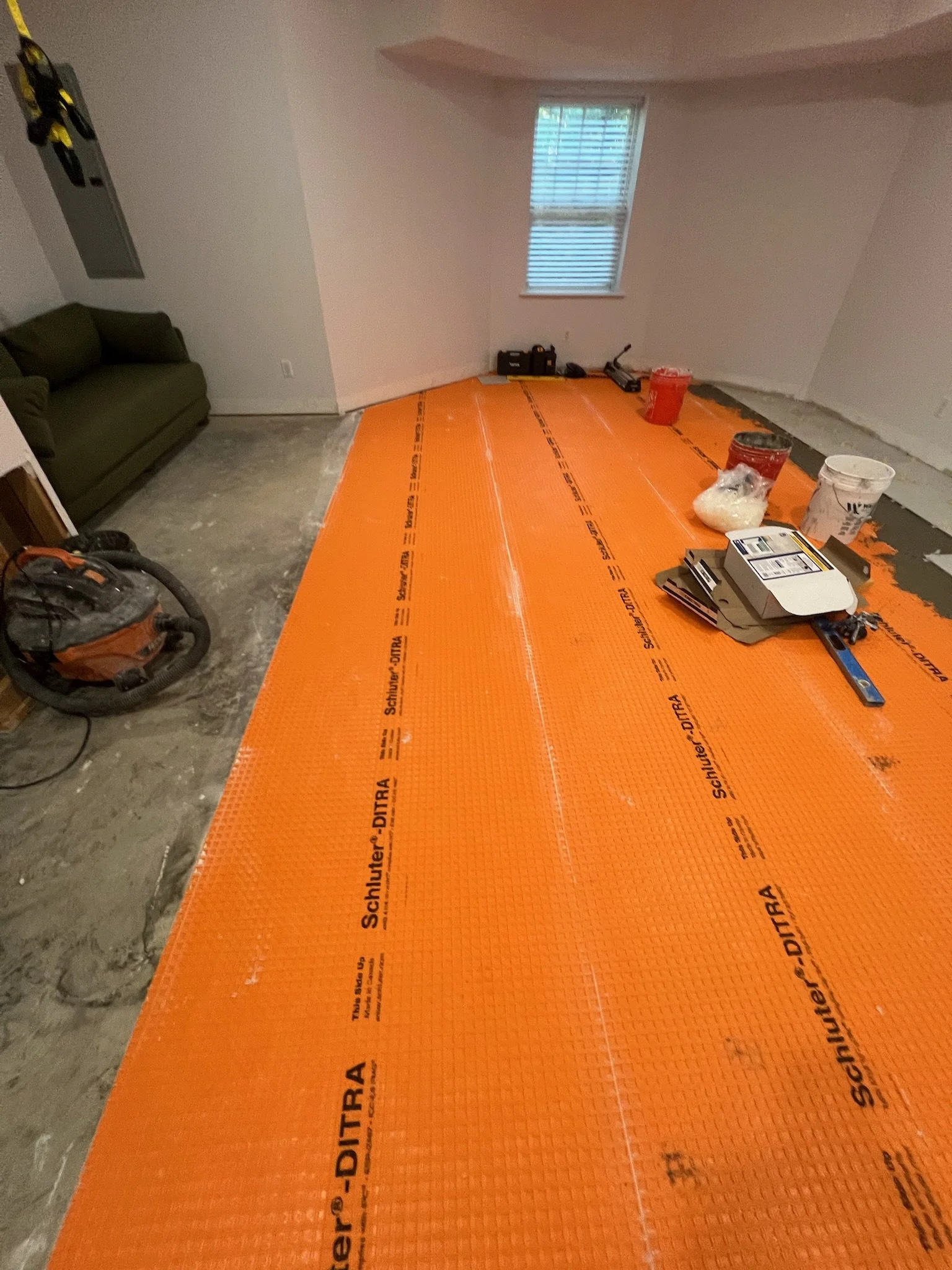 Room under construction with orange underlayment on the floor, tools, buckets, and a window in the background.