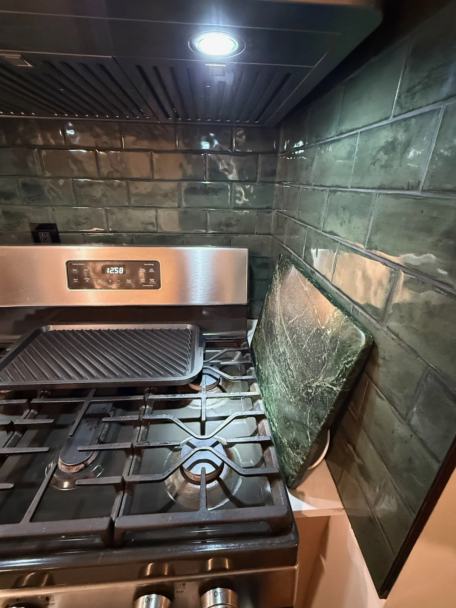 A kitchen stove with a grilling pan on the left and a green, weathered cutting board resting against the backsplash on the right, with dark green tiles and a black range hood above.