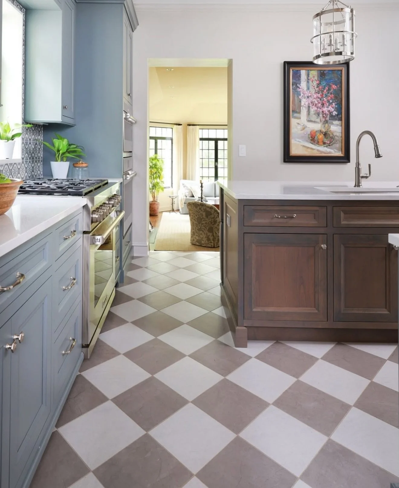 A kitchen with blue and brown cabinets, a tile floor with a checkerboard pattern, and a view into a living room with large windows and a green potted plant.