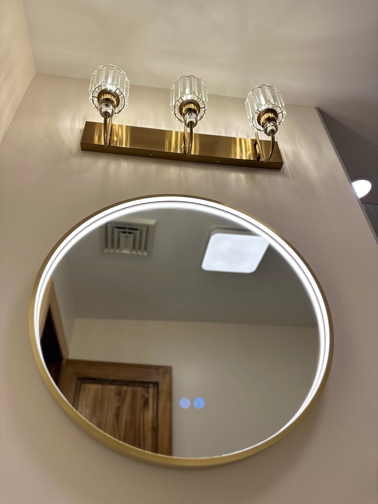 Close-up of a round mirror with a gold frame and a modern vanity light fixture with three clear glass bulb covers above it in a bathroom.