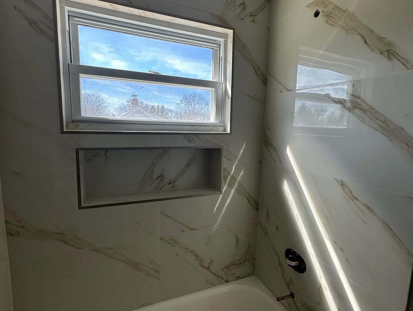 Bathroom with large marble tiles on the walls, a rectangular window showing a house and trees outside, and a built-in shelf in the wall. There is a fixture for a shower or faucet on the wall.