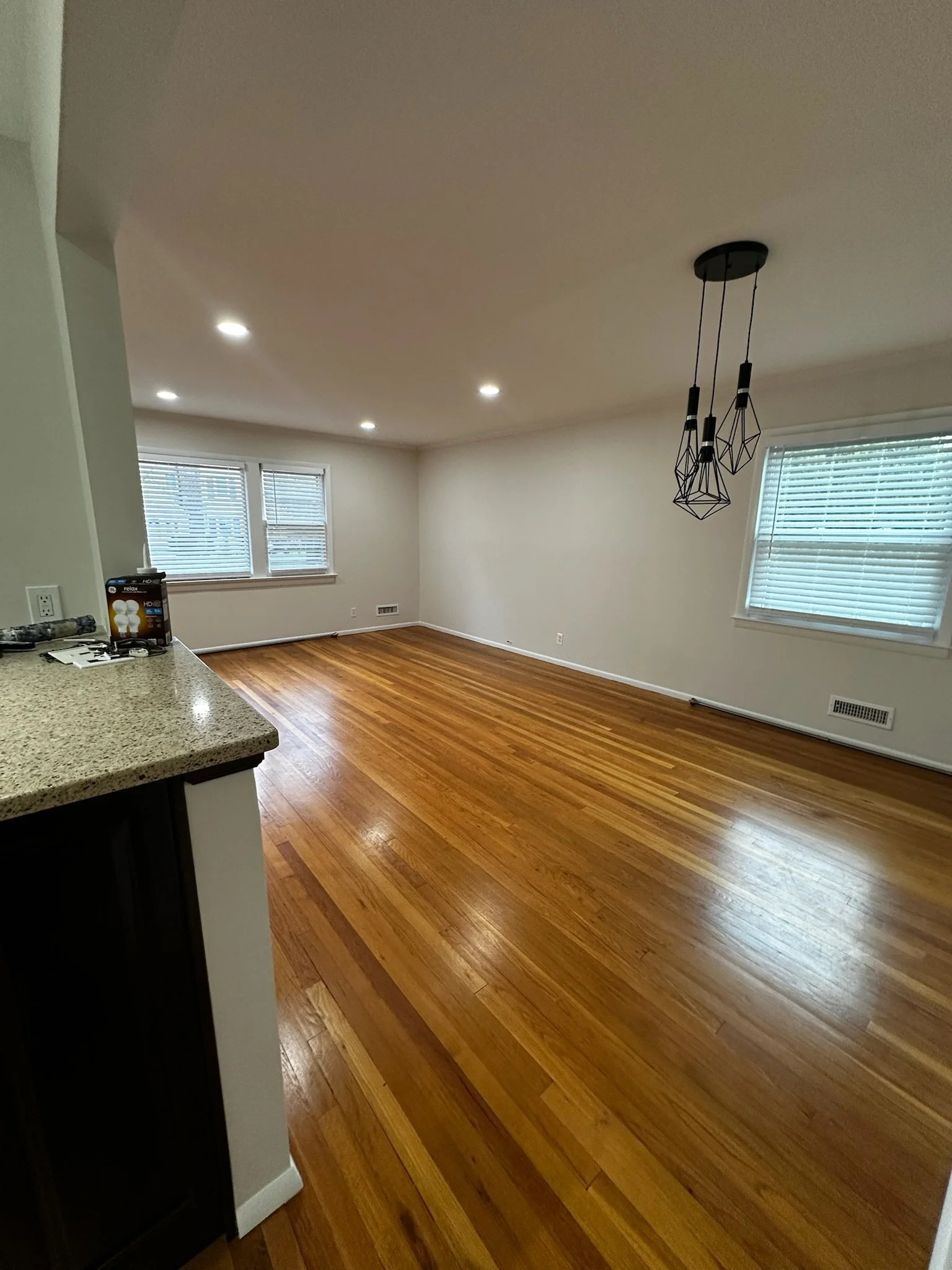 Empty living room with hardwood floors, white walls, three windows with blinds, and a modern black pendant light fixture.