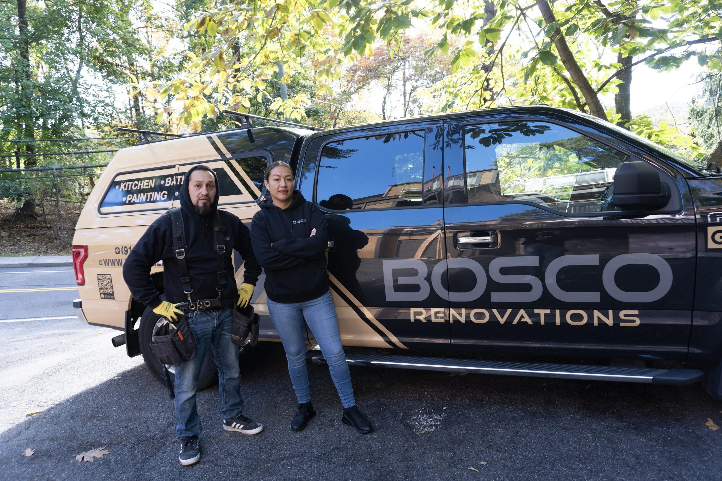 Two people, a man and a woman, standing next to a large black and beige Bosch Renovations vehicle parked on the side of a road surrounded by trees with green leaves. The man is wearing a black hoodie, jeans, sneakers, and yellow work gloves. The woman is wearing a black hoodie, blue jeans, and black boots. The vehicle has decals advertising kitchen, bath, and painting services.