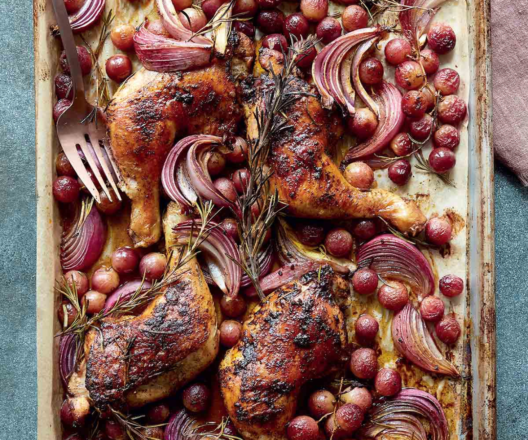 Roasted chicken thighs with red grapes, red onion slices, and sprigs of rosemary in a baking dish.