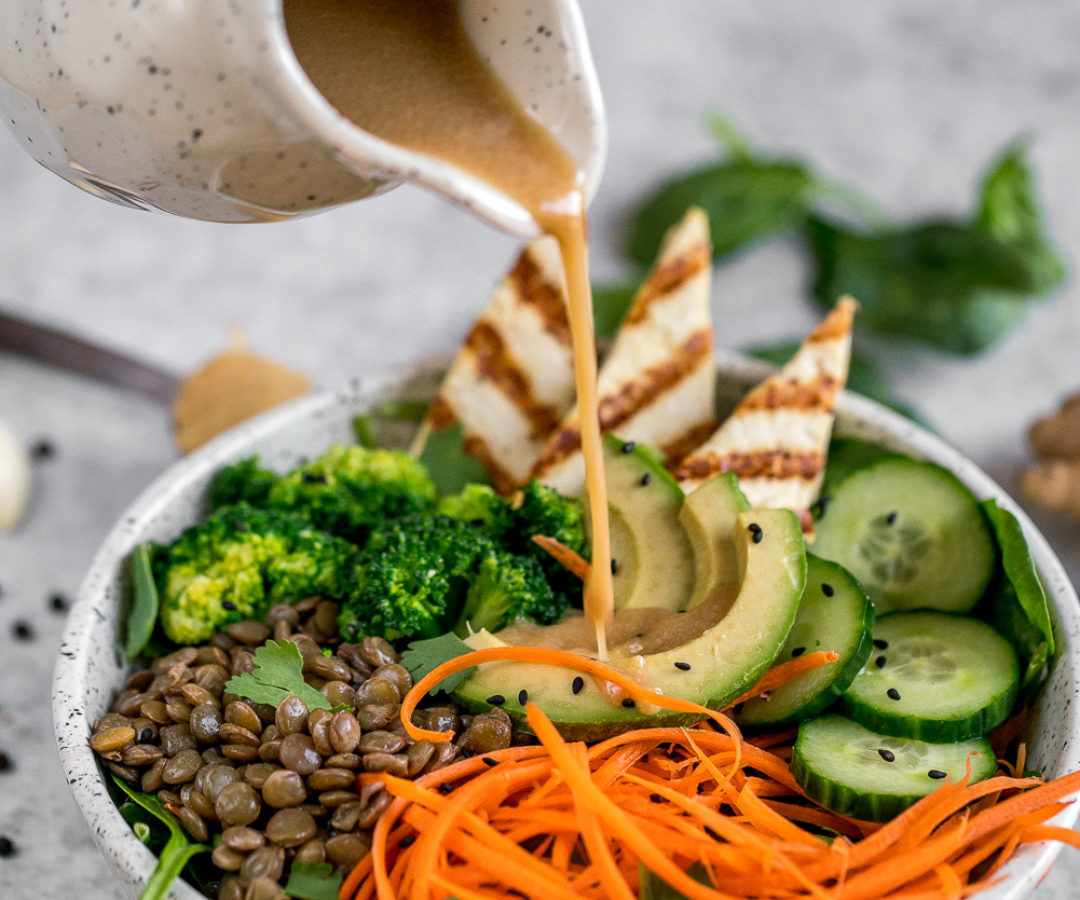A bowl of salad with broccoli, lentils, shredded carrots, cucumber slices, avocado, and grilled chicken strips, with dressing being poured over it.