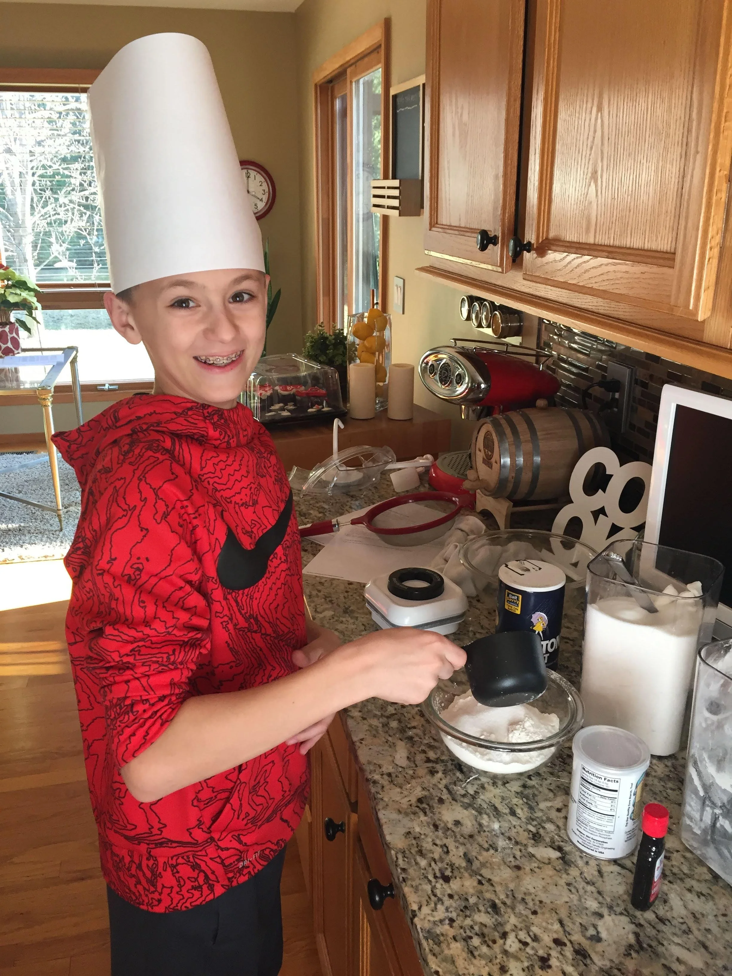 A young boy wearing a white chef’s hat and a red hoodie with a black Nike logo, smiling while mixing ingredients in a glass bowl in a kitchen.