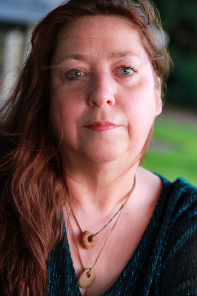 Close-up of a middle-aged woman with red hair, blue eyes, and fair skin, wearing a black top and two necklaces with wooden pendants, outdoors with blurred greenery in the background.