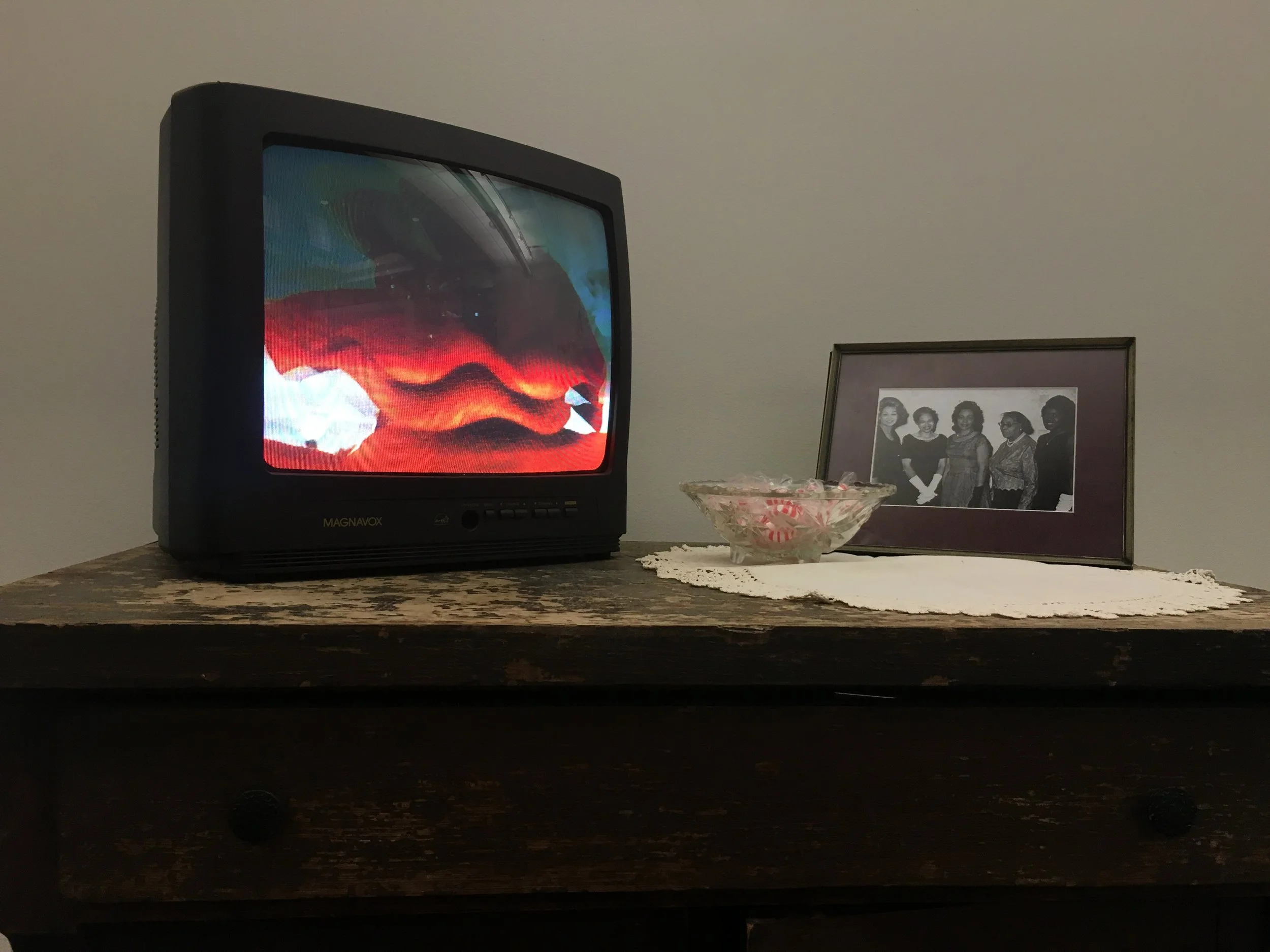 faith of our mothers (2016). On a small tube TV placed on side table, a red hat traces time leaving echoes of itself as the woman wearing it rocks her head side to side during a church service. Next to the TV is a bowl of peppermints and a photograph