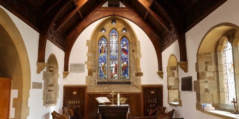 Interior of a church with wooden ceiling, stained glass windows, stone accents, and an altar with a cross and candles.