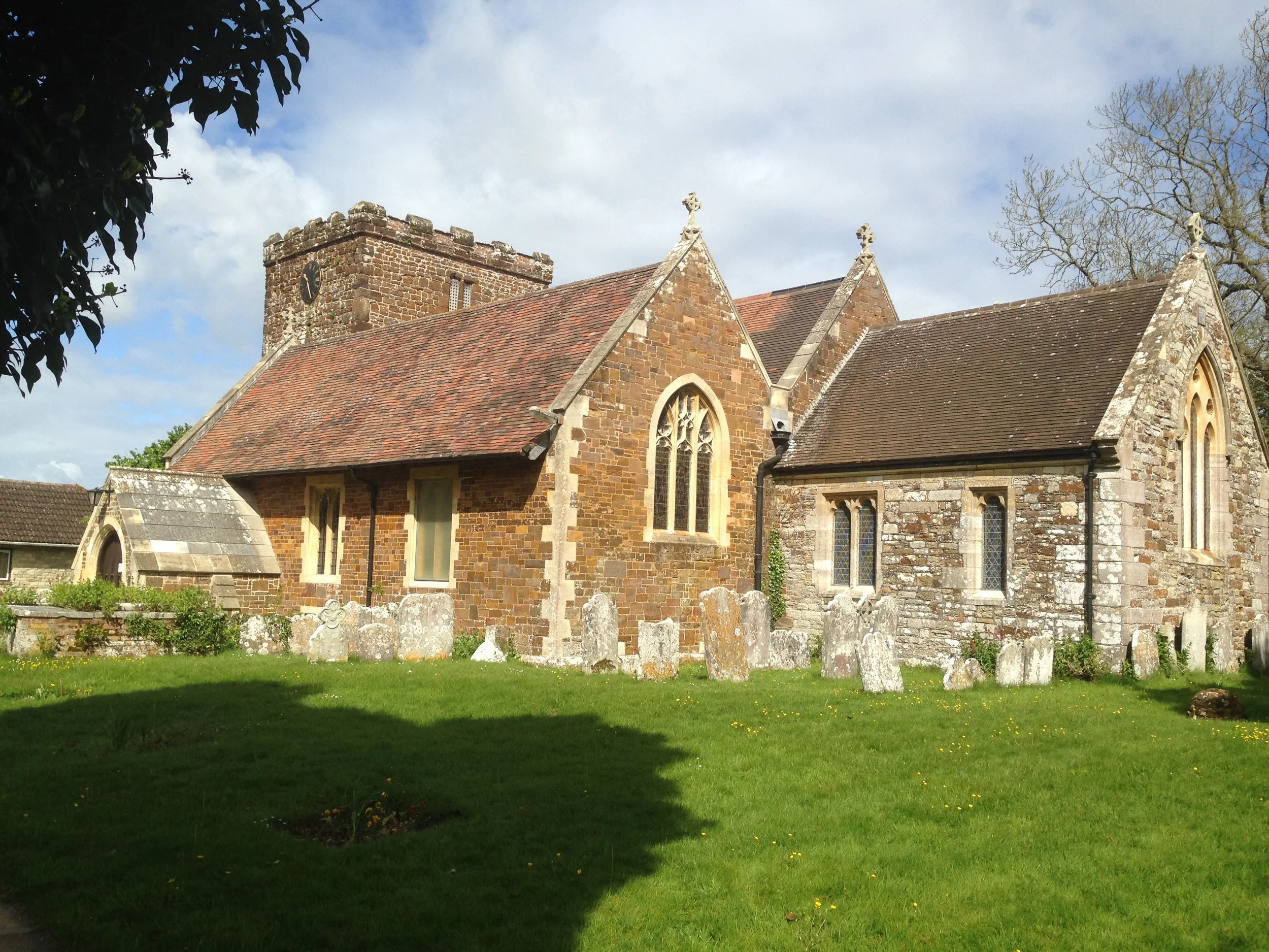 A historic stone church with a clock tower, surrounded by a grassy yard and old gravestones, under a partly cloudy sky.